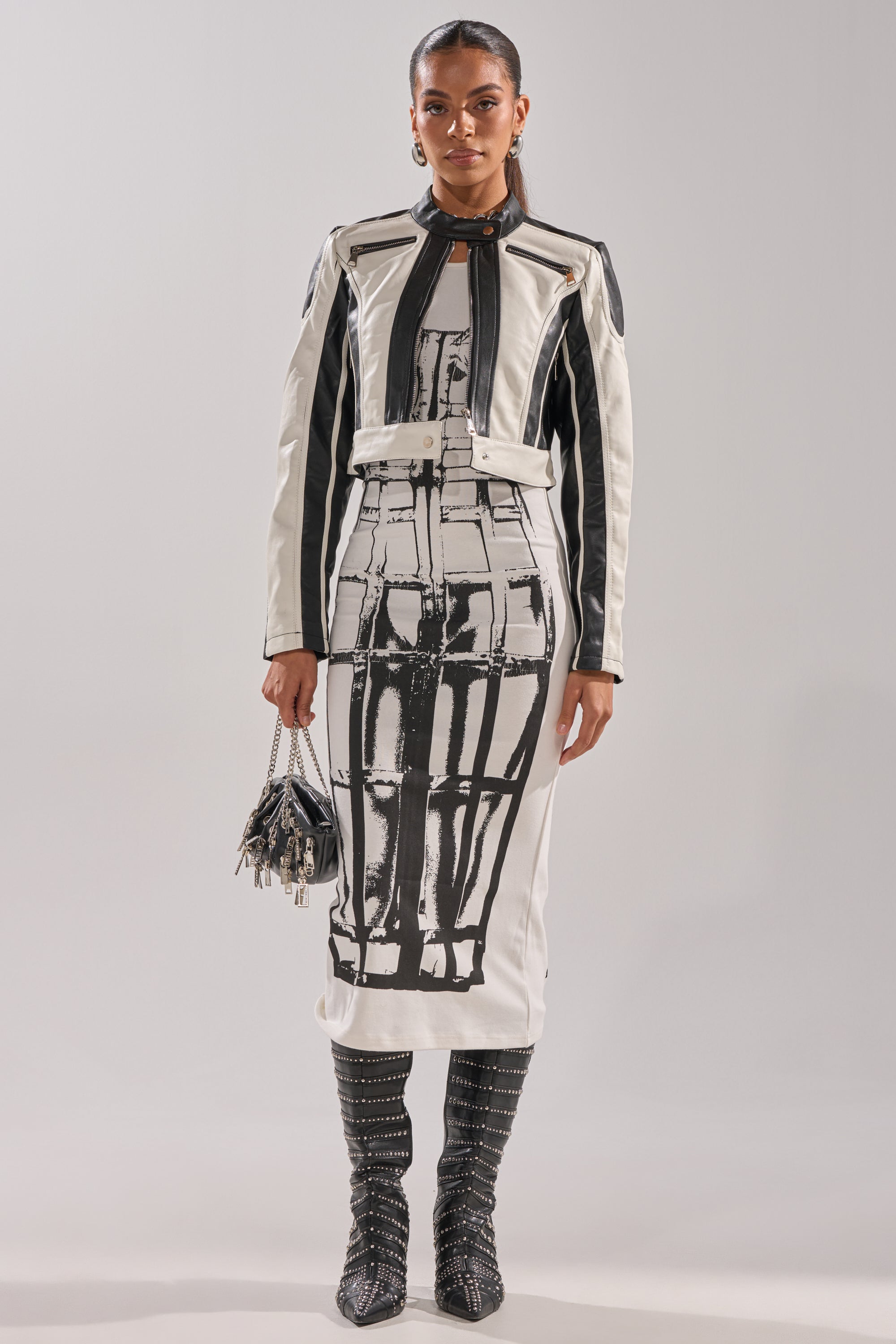 A woman wears the BETTER LUCK CROPPED MOTO IN BLACK WHITE jacket paired with a matching abstract print midi dress, black studded knee-high boots, and a small fringed handbag against a plain background. 7
