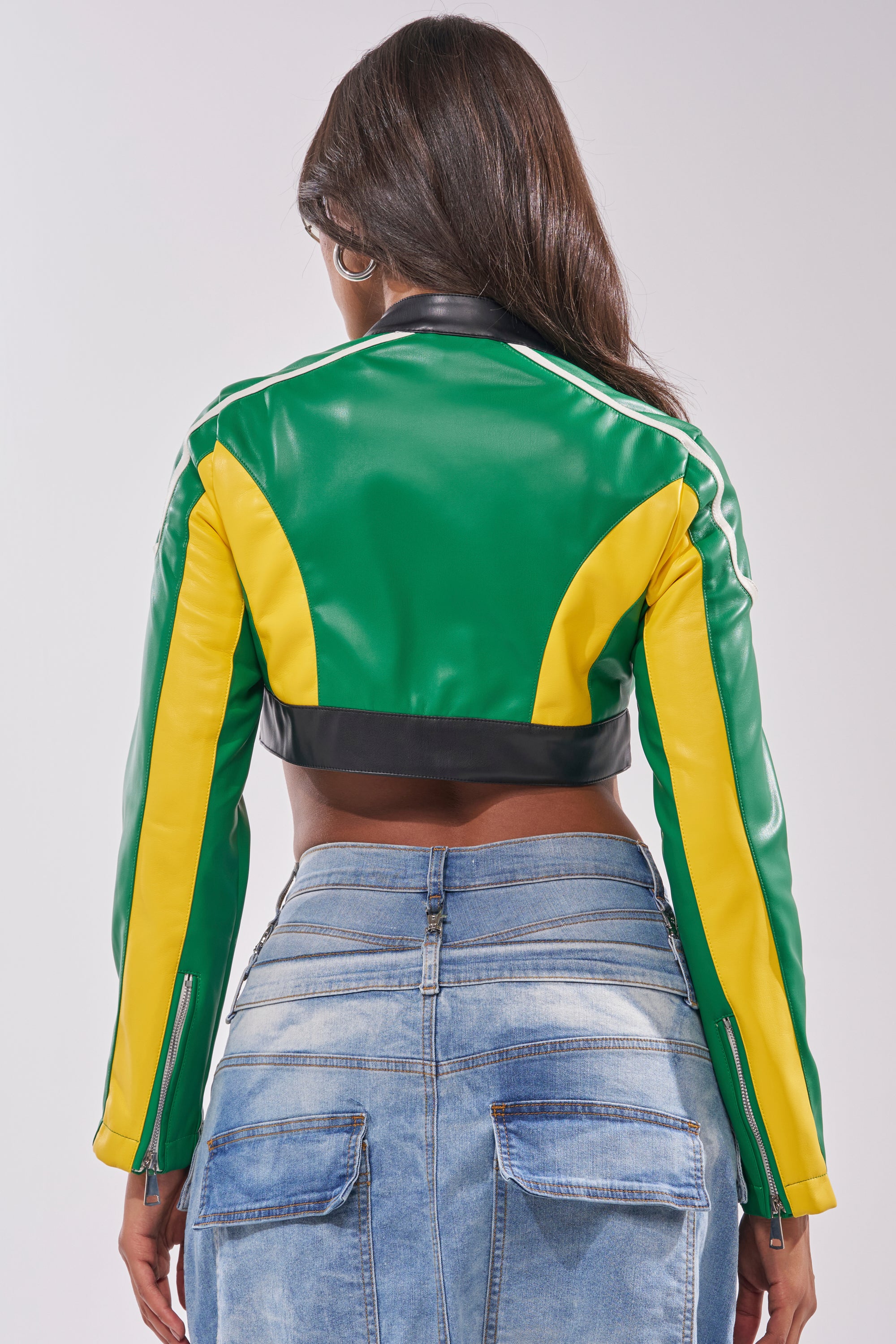 A person with long dark hair is shown from the back wearing the BIG FAN CROPPED MOTO—a bold green, yellow, and black streetwear jacket—paired with denim jeans against a plain light background. 4