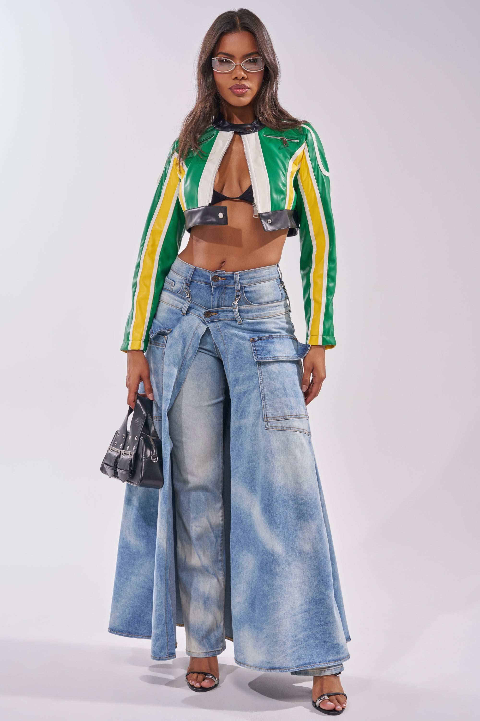 A woman in the BIG FAN CROPPED MOTO jacket, glasses, wide-leg light blue jeans, black sandals, and a small black handbag poses confidently against a plain backdrop. 5