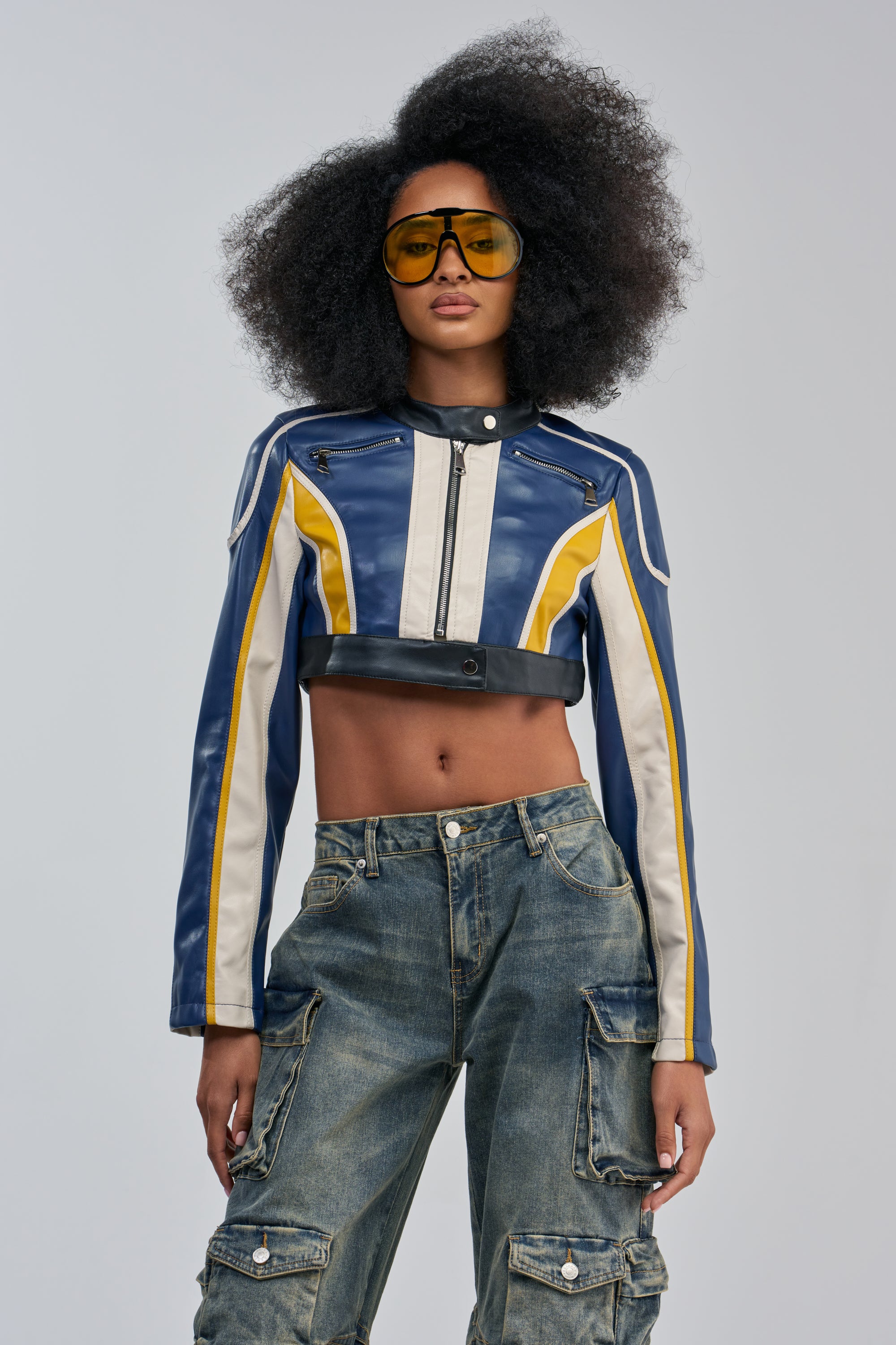 A woman with voluminous curly hair stands against a plain light gray background, wearing yellow-tinted sunglasses, cargo jeans, and the FAST LANE RACER MOTO blue and yellow colorblocked jacket.