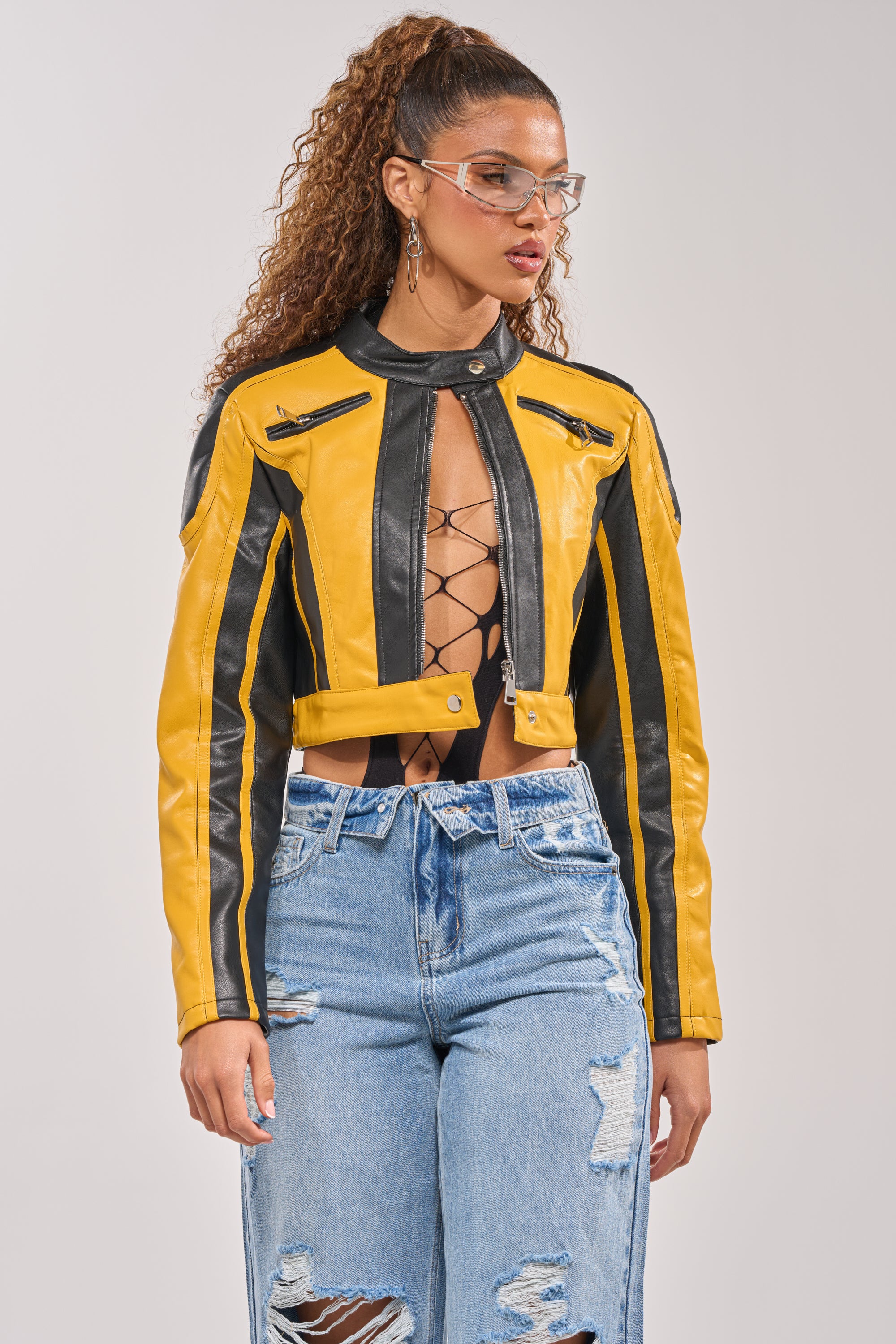A woman wears the ON YOUR MARKS RACER MOTO IN YELLOW MULTI jacket, a cropped silhouette paired with a lace-up black top and ripped high-waisted blue jeans. She has curly hair in a ponytail and clear fashion glasses for a bold biker look. 0