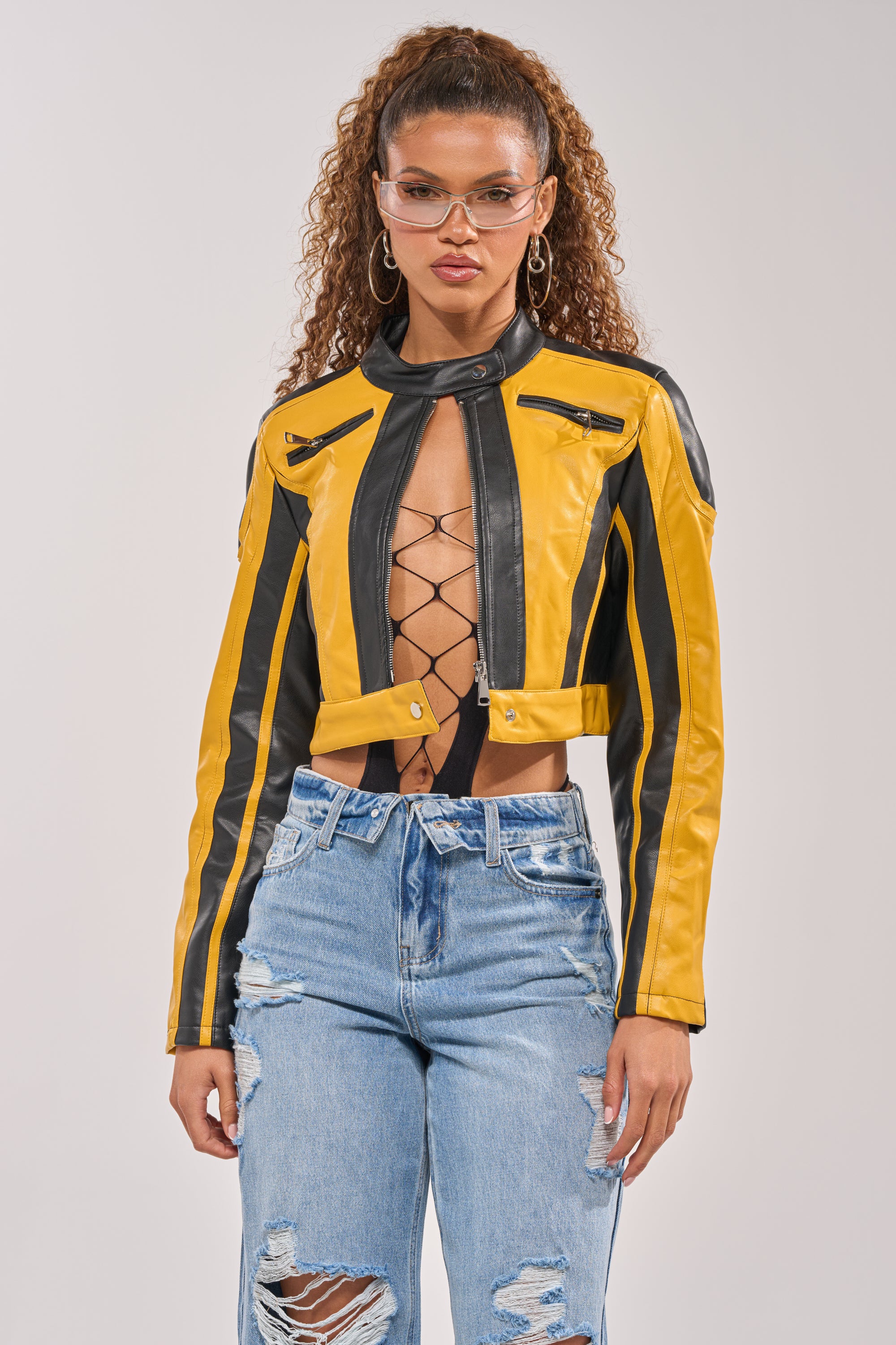 A woman with curly hair and clear glasses wears the ON YOUR MARKS RACER MOTO IN YELLOW MULTI jacket, a black lace-up top, and ripped blue jeans, standing against a plain light background. 1
