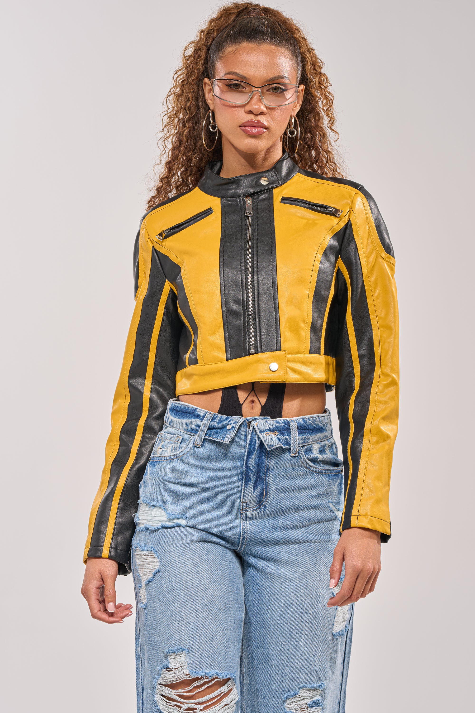 A woman models the ON YOUR MARKS RACER MOTO IN YELLOW MULTI jacket with vertical stripes and high-waisted ripped blue jeans, standing against a light gray background for a bold biker look. 2