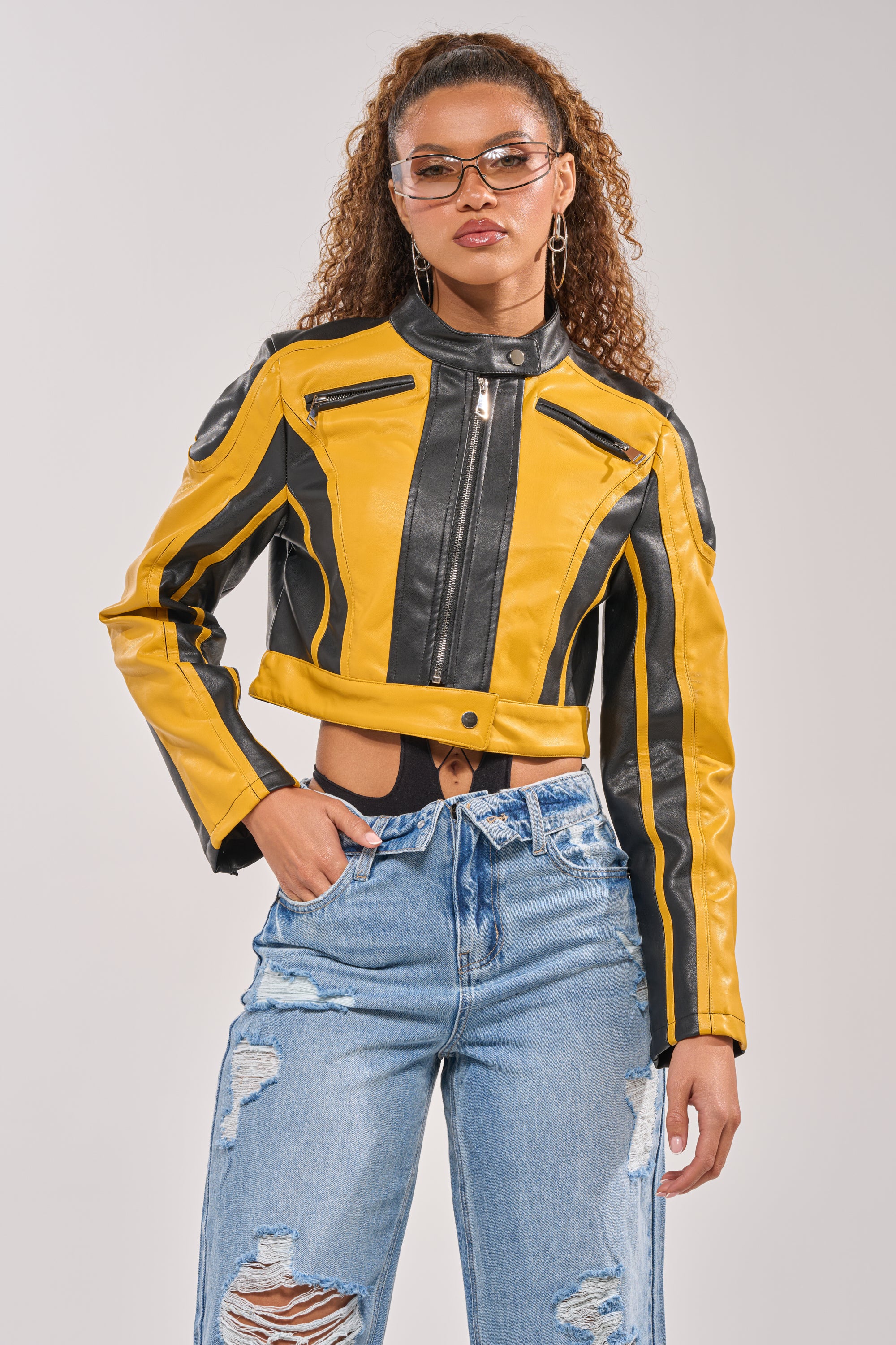 A woman with curly hair confidently poses in the ON YOUR MARKS RACER MOTO IN YELLOW MULTI jacket, paired with ripped blue jeans, hoop earrings, and clear glasses, one hand in her pocket against a plain background. 3