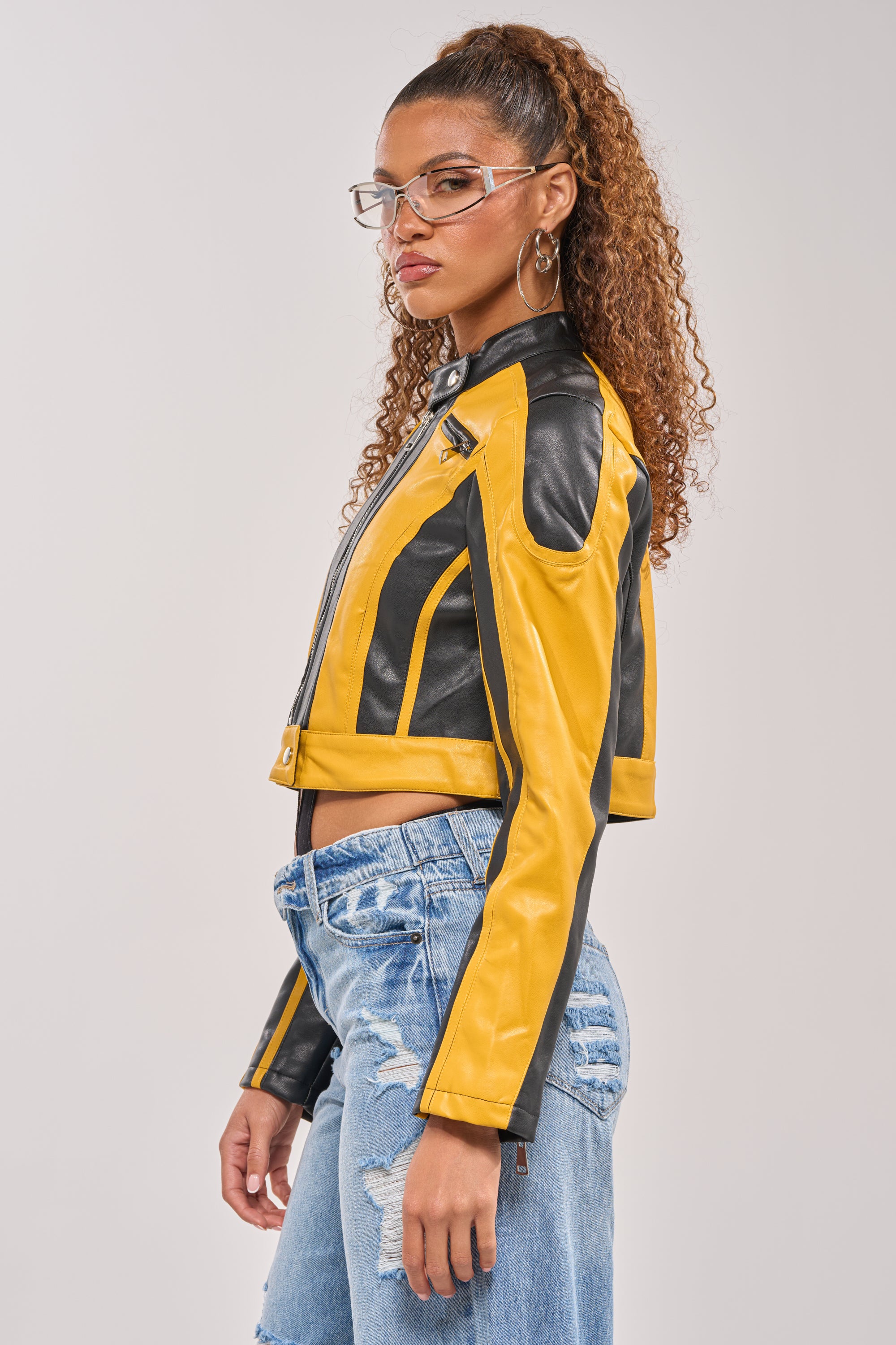 A woman with curly hair in a high ponytail wears clear glasses, hoop earrings, ripped blue jeans, and the ON YOUR MARKS RACER MOTO IN YELLOW MULTI jacket, standing in profile against a plain background. 4