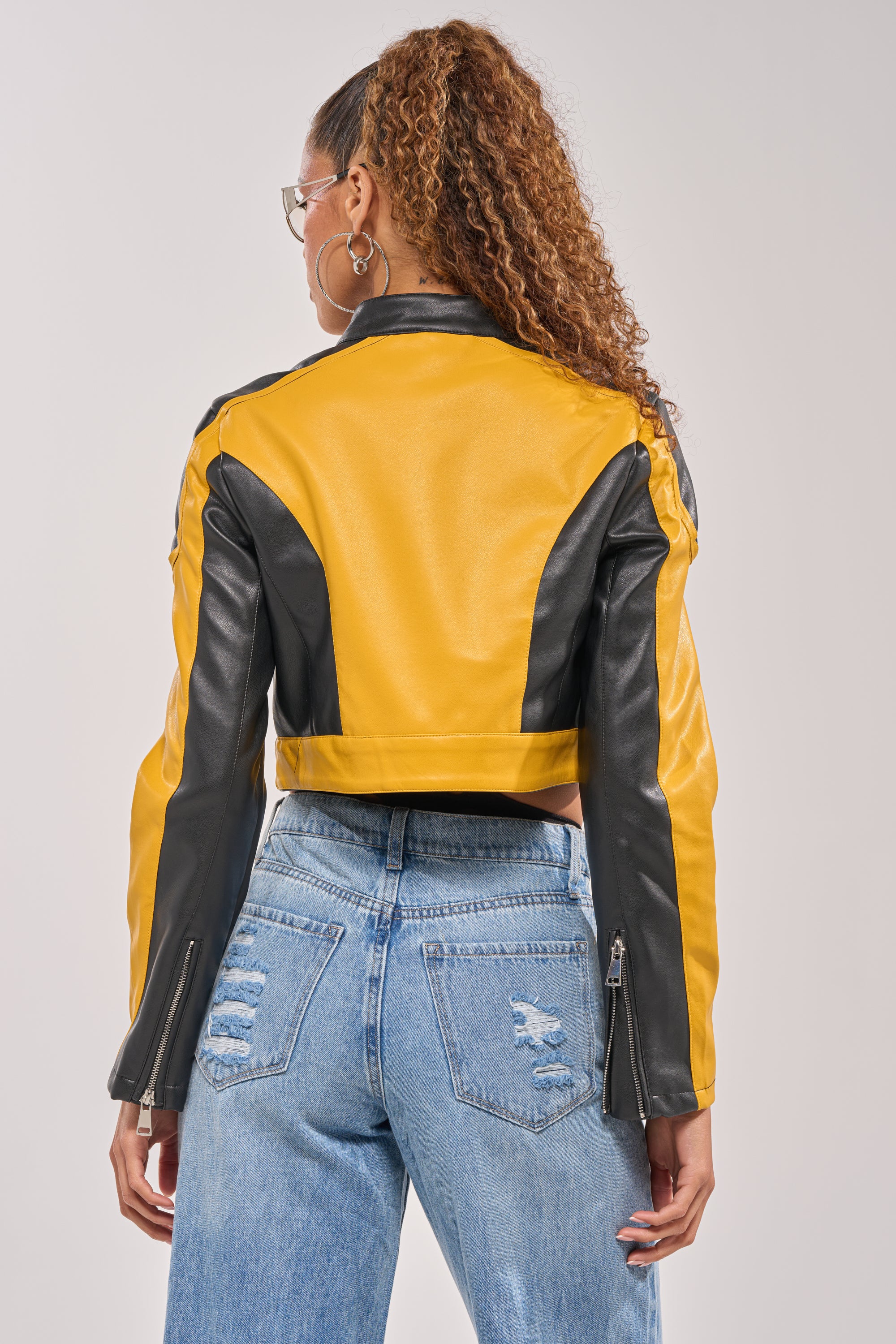 A woman with curly hair in a high ponytail, shown from the back, wears the ON YOUR MARKS RACER MOTO IN YELLOW MULTI—a cropped yellow and black racing-inspired jacket—with light blue ripped jeans against a plain light background. 5