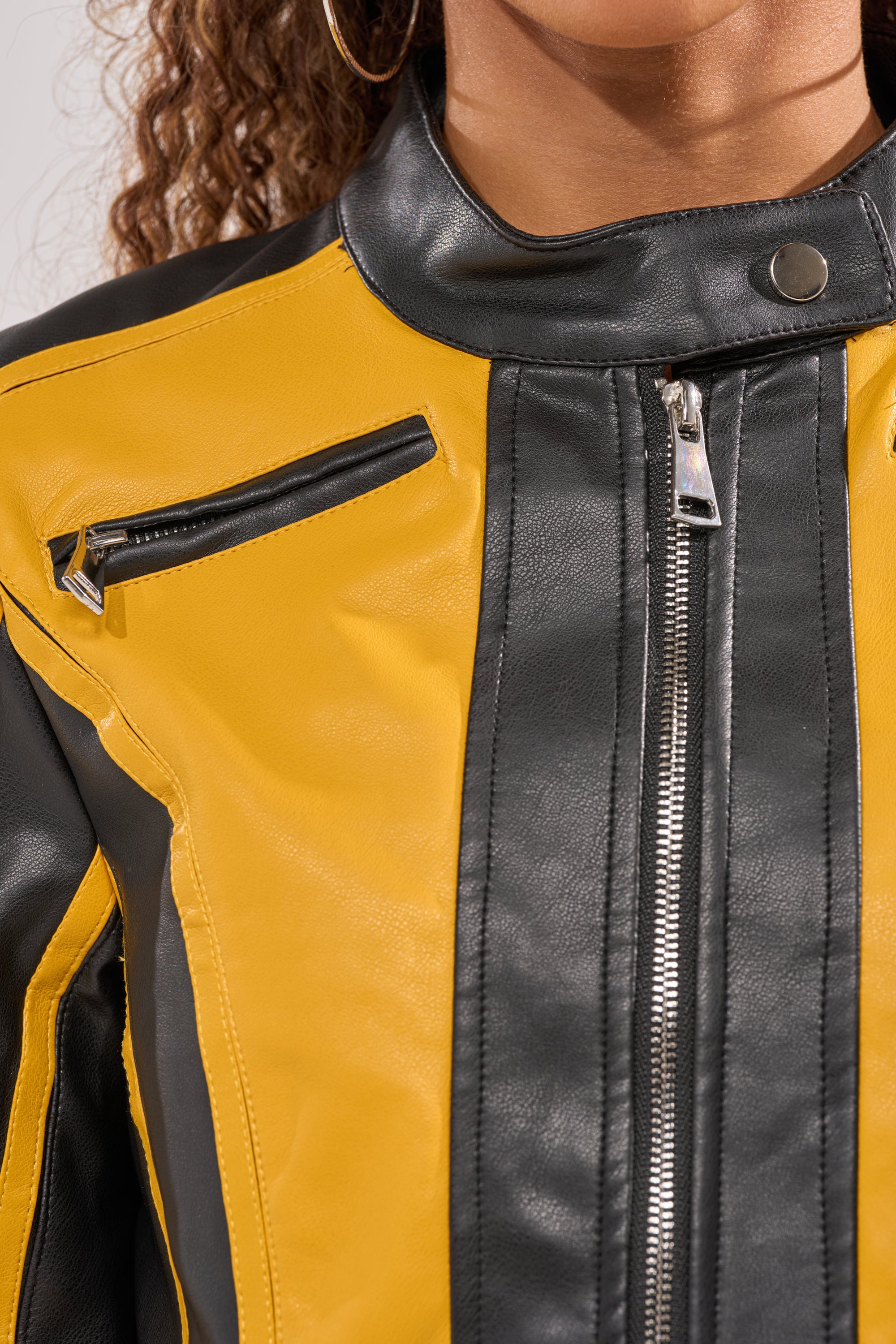 Close-up of a cropped silhouette in the ON YOUR MARKS RACER MOTO IN YELLOW MULTI, featuring racing-inspired yellow and black panels, a vertical zipper, snap collar, and angled chest pocket. Only the lower face and curly hair are visible. 8