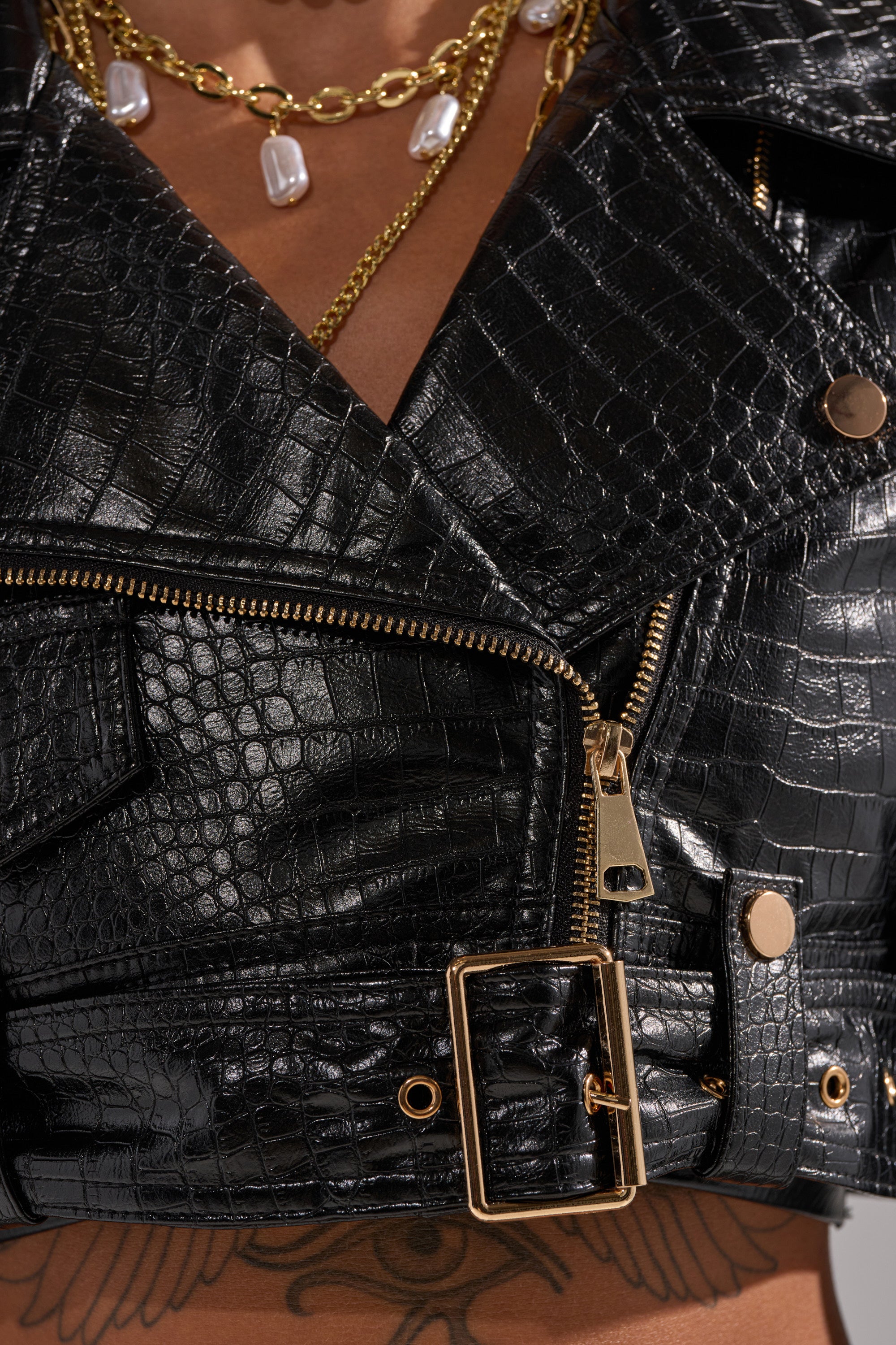 Close-up of a person wearing the EVERYDAY MOTO IN BLACK CROC jacket with gold zippers and buckles, layered gold necklaces with pearl accents, and a visible tattoo on their lower abdomen. 8