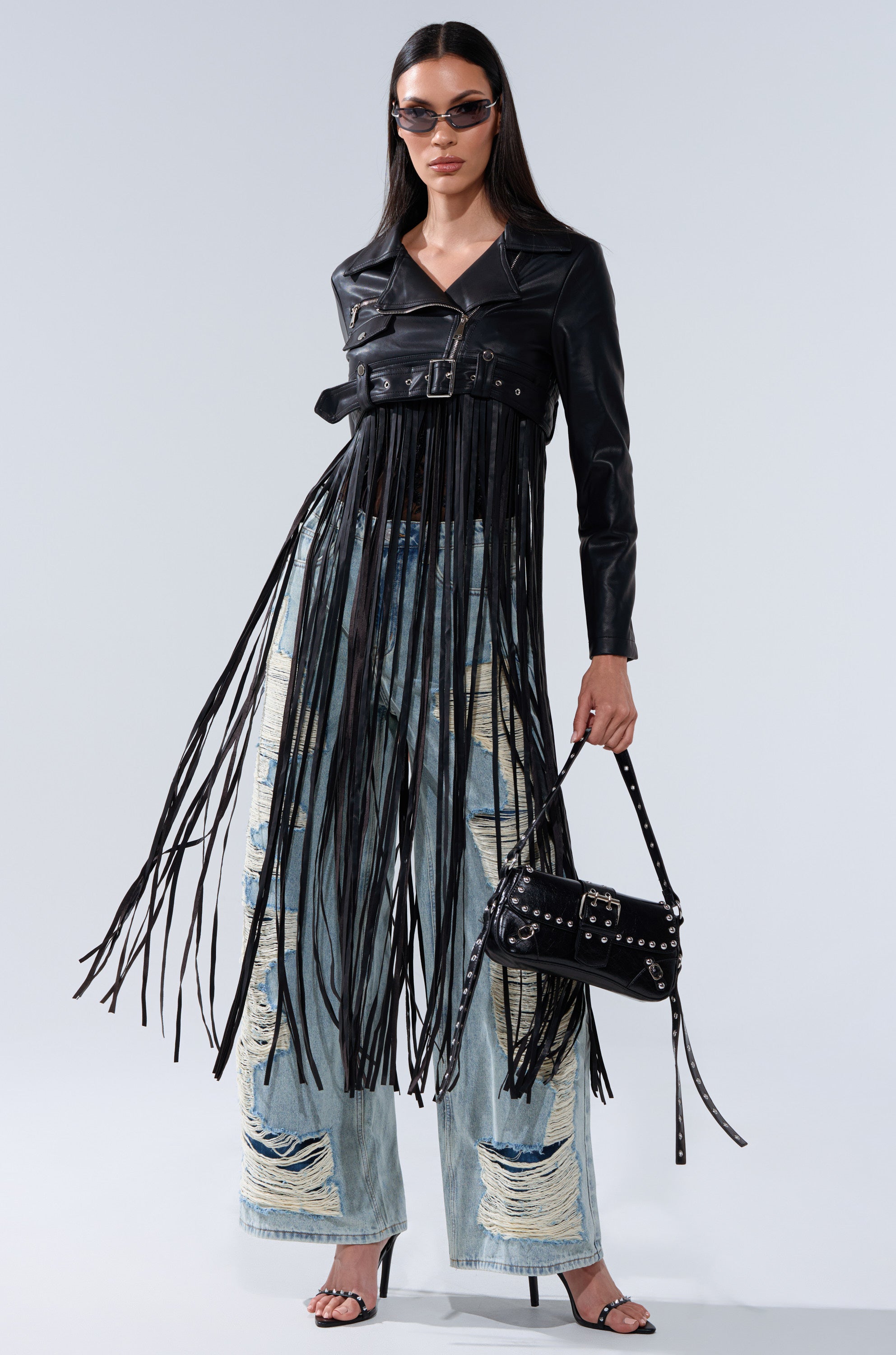 A woman poses confidently in the SWING MY WAY FRINGE MOTO jacket, distressed wide-leg jeans, black heels, dark sunglasses, and a studded black handbag against a plain light background.