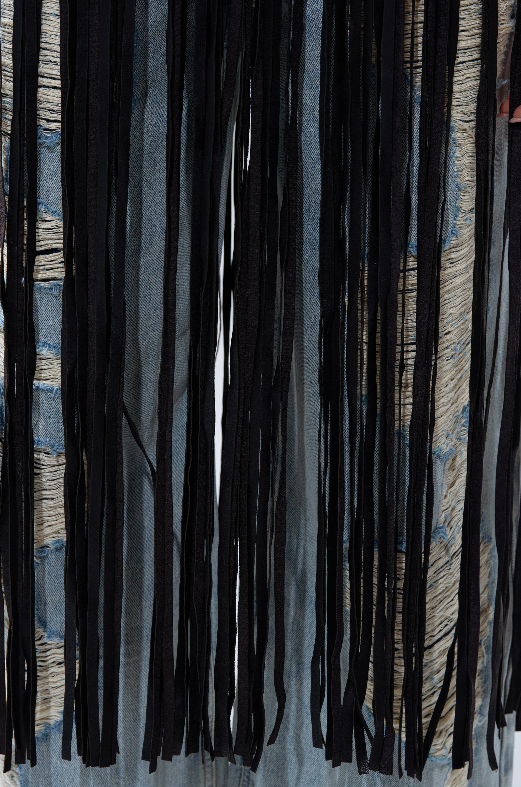 Close-up of distressed blue denim jeans with frayed holes, partially covered by the vertical black fringe strips from the cropped SWING MY WAY FRINGE MOTO jacket.