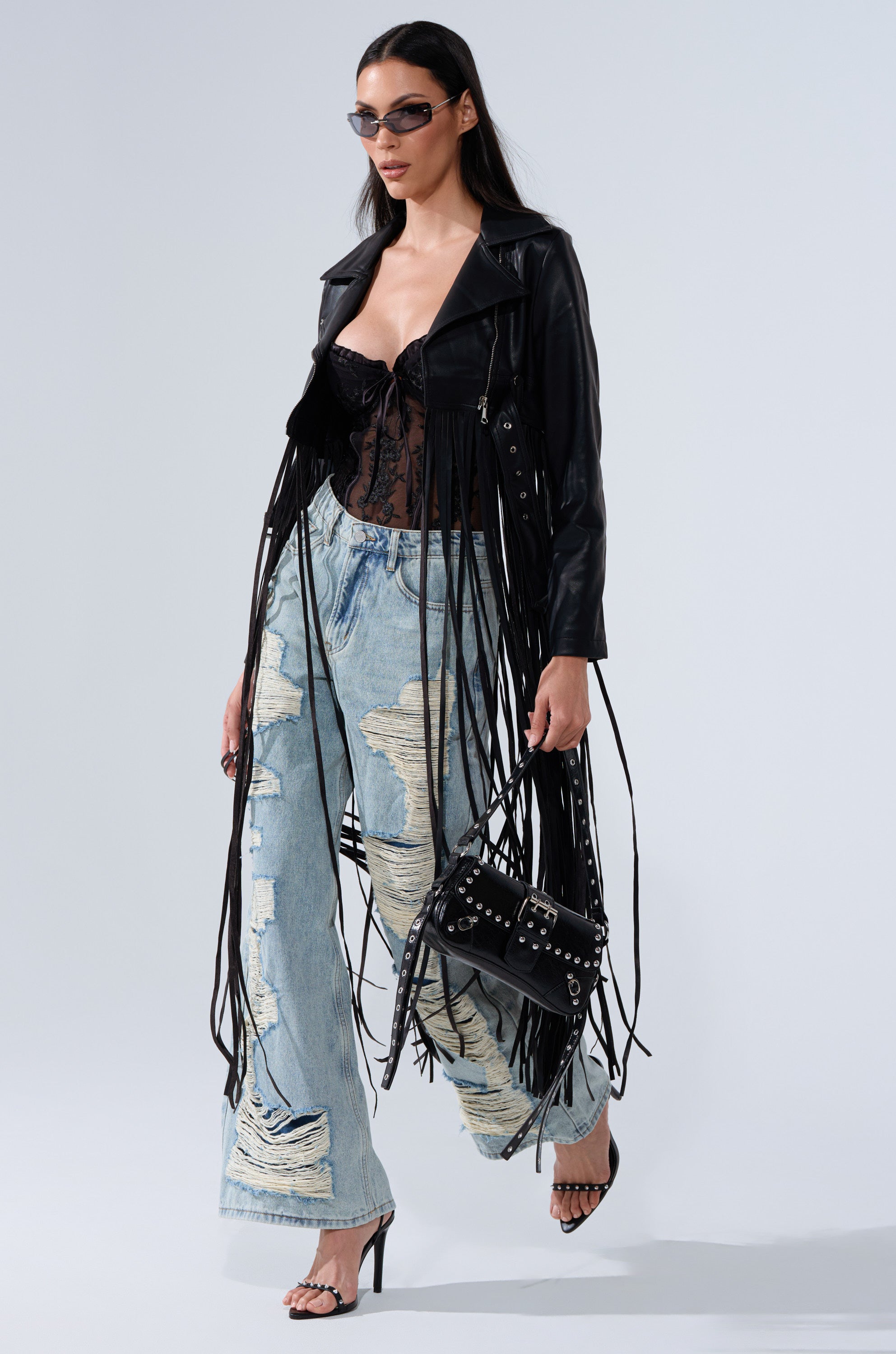 A woman walks confidently in sunglasses, the SWING MY WAY FRINGE MOTO jacket, a sheer lace top, distressed wide-leg jeans, heeled sandals, and carries a black studded handbag.