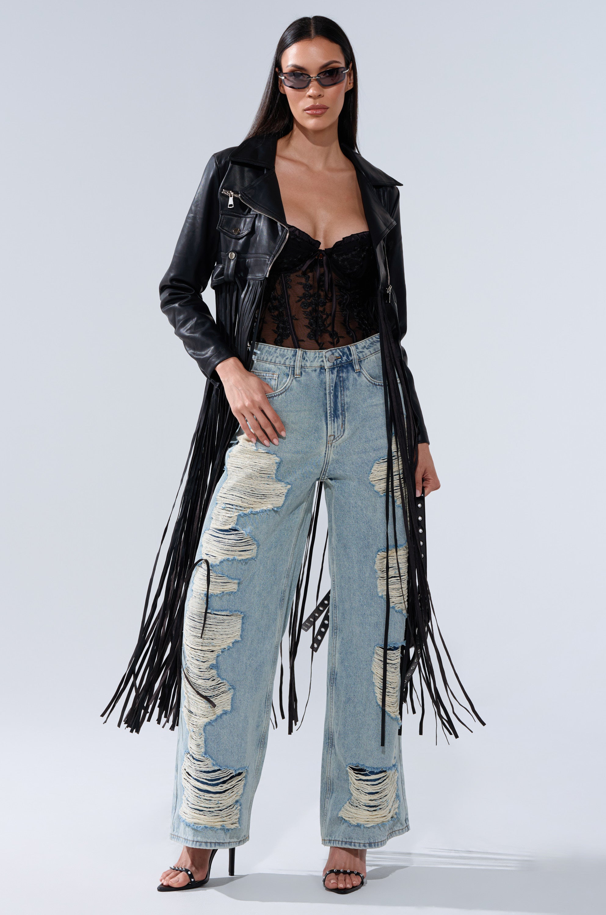 A woman poses confidently against a plain background wearing sunglasses and the SWING MY WAY FRINGE MOTO—a black fringed jacket—layered over a lace corset top, with light blue distressed wide-leg jeans and black heeled sandals.
