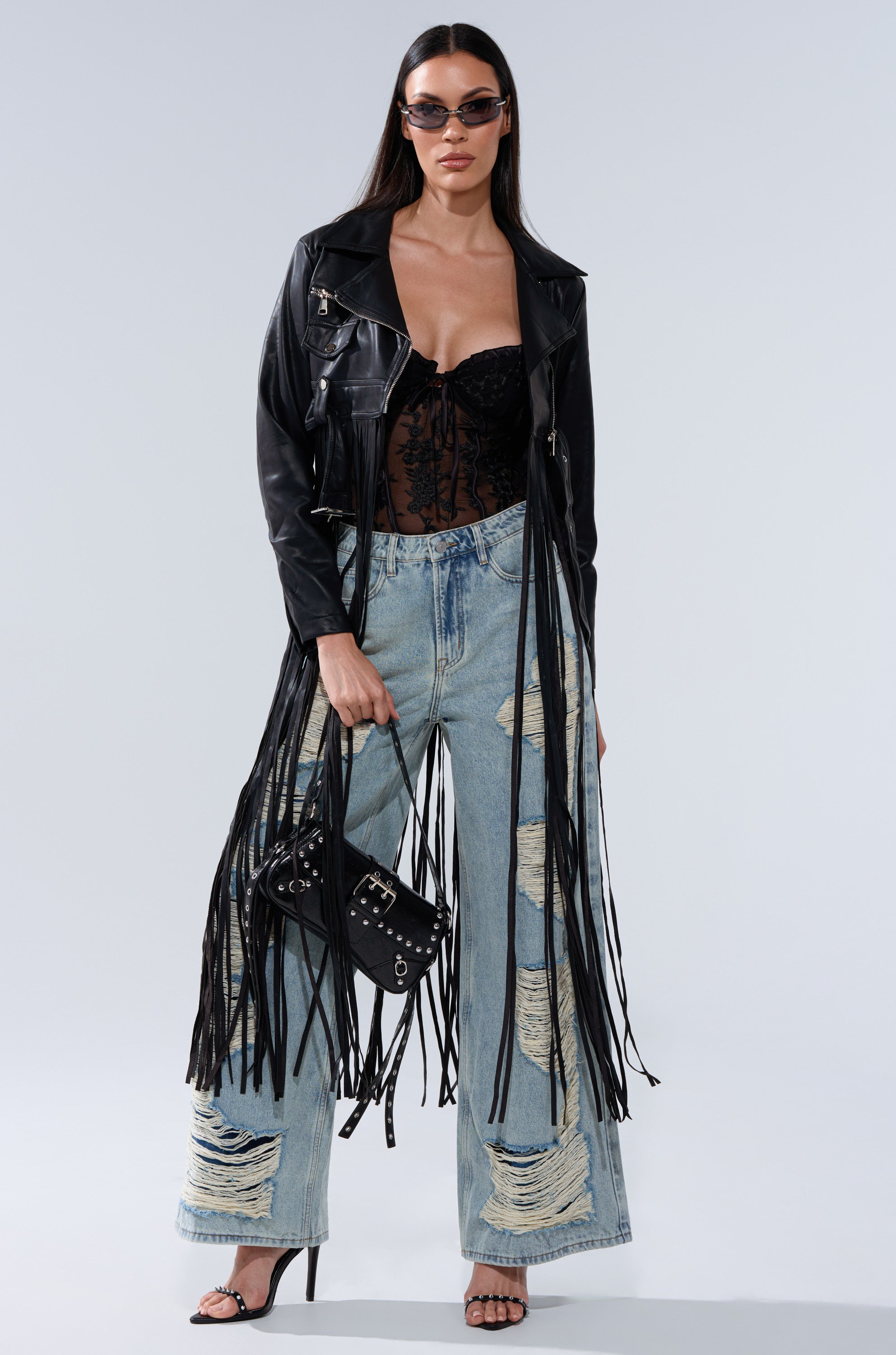 A woman wearing sunglasses, a black lace top, the SWING MY WAY FRINGE MOTO jacket, ripped wide-leg jeans, black high heels, and holding a black handbag with metal details.