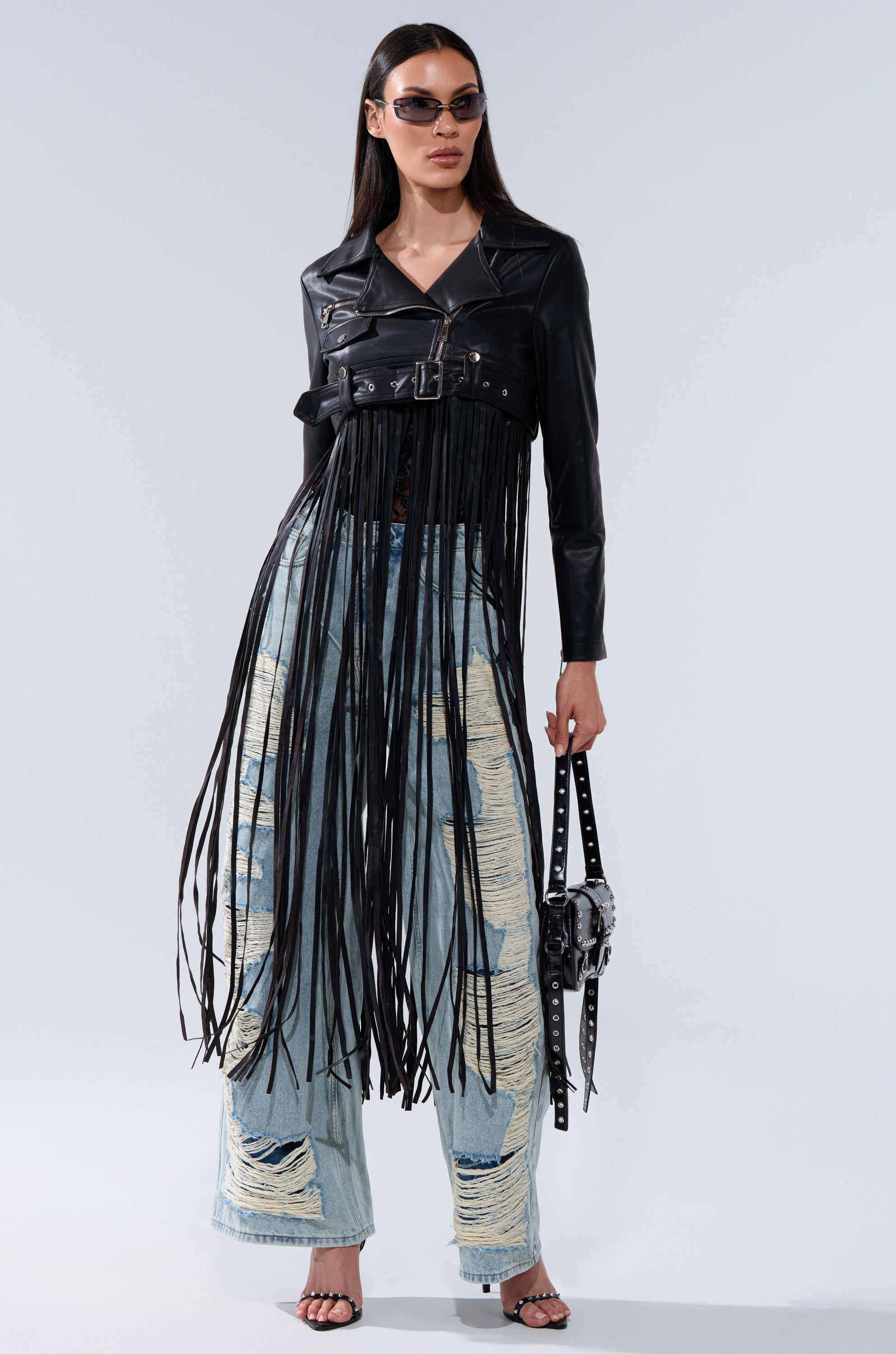 A woman stands confidently against a plain background, wearing sunglasses, the SWING MY WAY FRINGE MOTO jacket with long fringe details, distressed light blue jeans, black heeled sandals, and holding a studded belt and small bag.