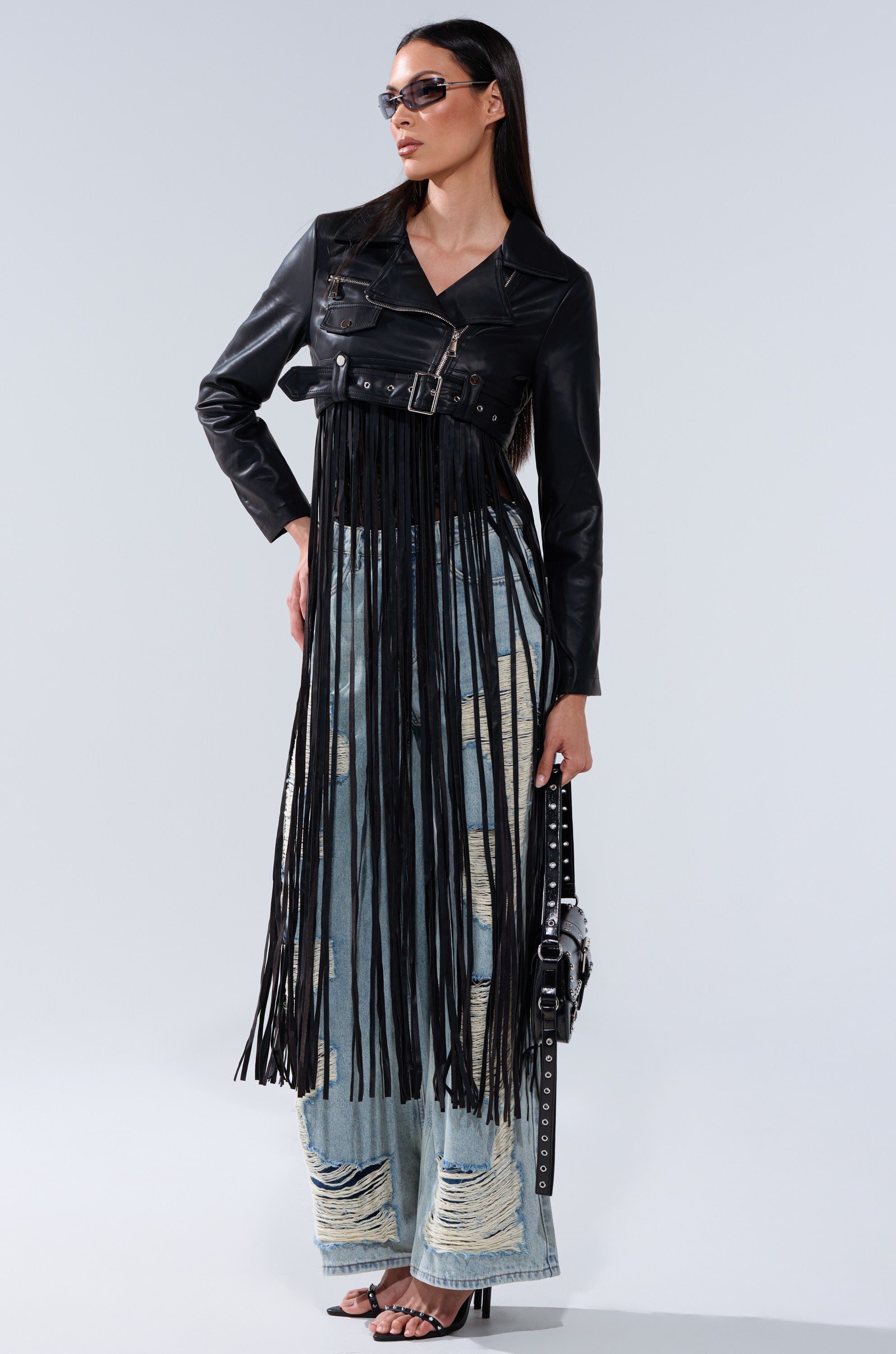 A woman wears sunglasses, the SWING MY WAY FRINGE MOTO black jacket with long fringe, distressed wide-leg jeans, heeled sandals, and holds a black studded handbag against a plain background.