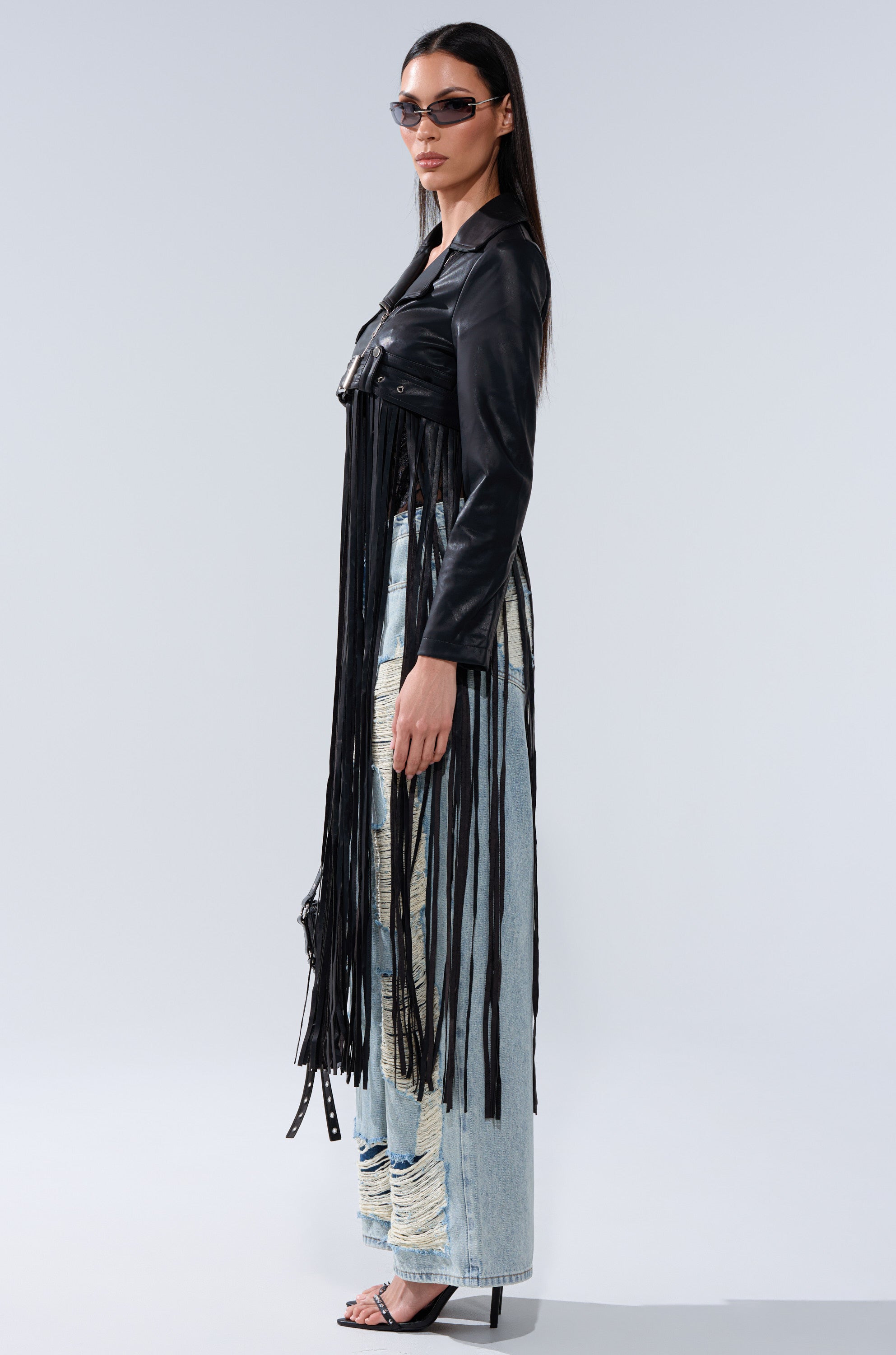 Wearing the SWING MY WAY FRINGE MOTO faux leather jacket, a woman in sunglasses pairs it with distressed light blue jeans and black heeled sandals, posing sideways against a plain light background.