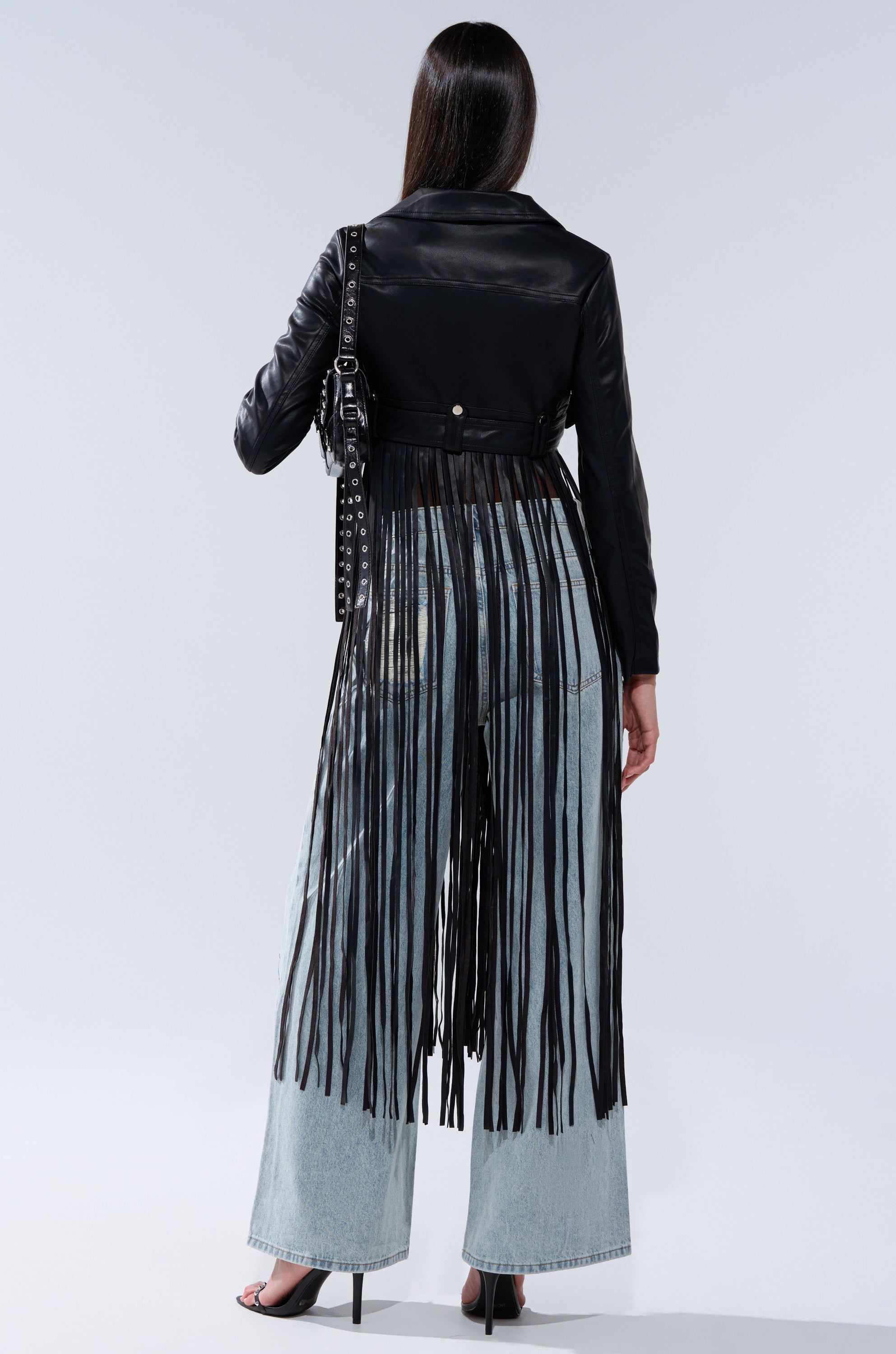 A person stands facing away, wearing the SWING MY WAY FRINGE MOTO jacket with long black fringe over light blue wide-leg jeans and black heels, holding a black studded bag.