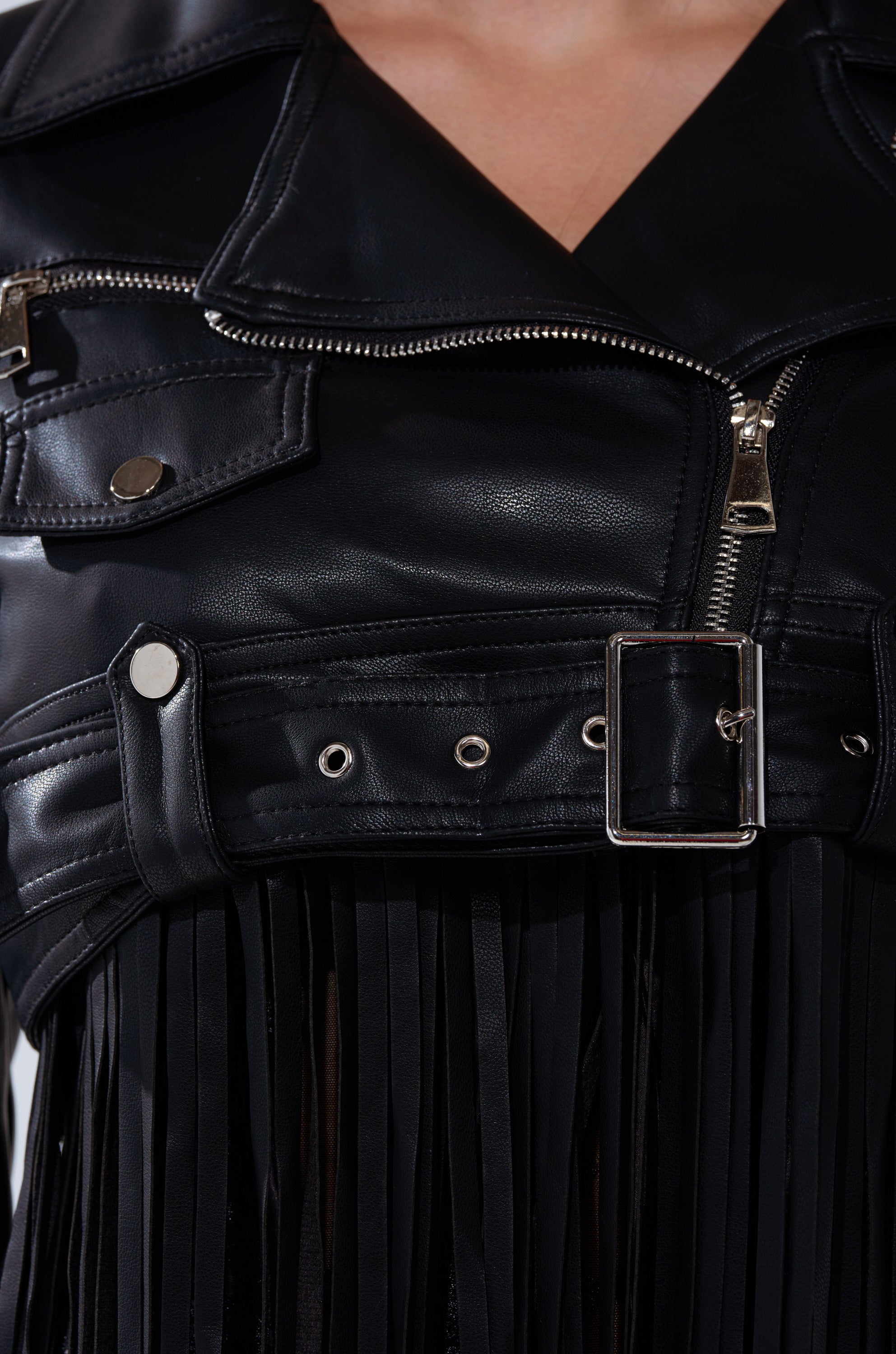 Close-up of a person wearing the SWING MY WAY FRINGE MOTO—a black faux leather cropped jacket with silver zipper, buckle, and button details, finished with stylish fringe accents along the lower edge.