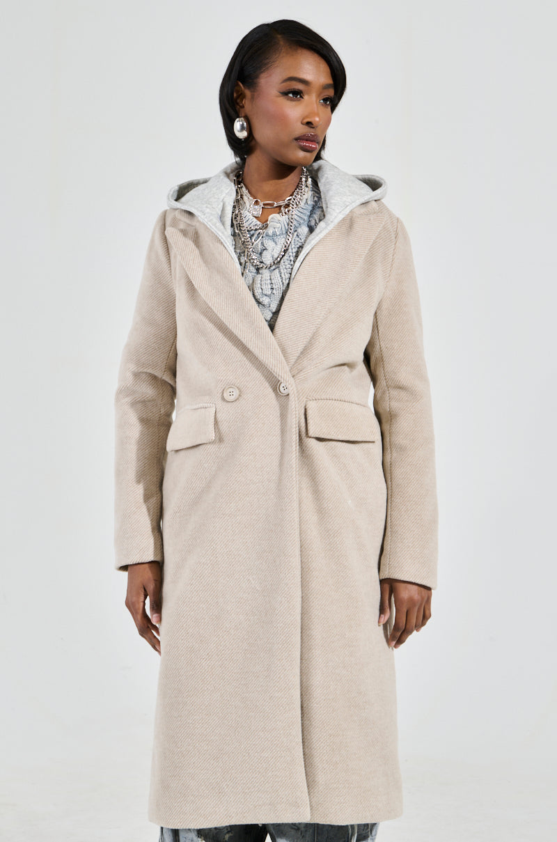 Dressed in the ONLY ONE LAYERED TRENCH, a long beige coat featuring an oversized silhouette, an individual wears it over a patterned top, accessorized with layered necklaces and hoop earrings. The ensemble is enhanced by lightweight makeup and a chic short hairstyle, all set against a plain gray background.