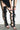 Wearing a black skirt, a person adjusts the AZALEA WANG KRISTINE BLACK STRAPPY UPPER GLADIATOR SANDAL—tall, thigh-high lace-up heels with intricate straps—indoors on a light floor.
