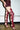 A person stands against a white wall and concrete floor, wearing a textured burgundy mini dress and AZALEA WANG KRISTINE BURGUNDY STRAPPY GLADIATOR SANDALS with multiple straps and stiletto heels.