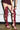 A person in a short maroon dress adjusts the AZALEA WANG KRISTINE BURGUNDY STRAPPY GLADIATOR SANDAL, featuring tall, strappy stiletto heels with multiple bows and open toes, wrapping up to the thighs. Their hands are visible on a concrete floor.