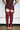 A person wearing a short burgundy dress with floral details and the AZALEA WANG KRISTINE BURGUNDY STRAPPY GLADIATOR SANDAL stands indoors against a white wall and window.