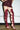 Someone stands indoors on a concrete floor wearing a short, textured burgundy dress and the AZALEA WANG KRISTINE BURGUNDY STRAPPY GLADIATOR SANDAL—knee-high heels with intricate lace-up details.