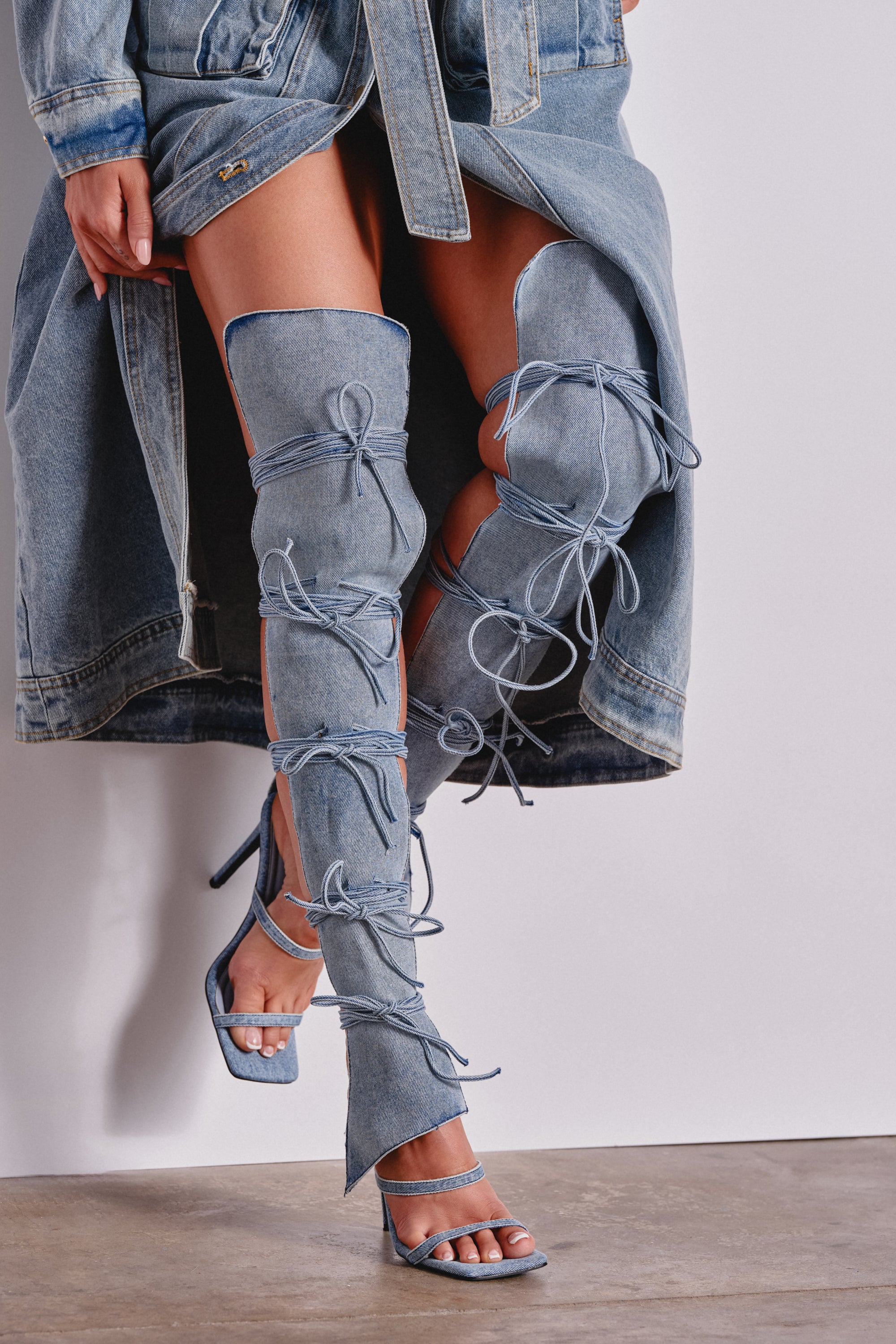 A person stands against a plain white wall and concrete floor, wearing a long denim coat and unique thigh-high stiletto boots tied with bows, paired with the AZALEA WANG KRISTINE DENIM STILETTO GLADIATOR SANDAL. 0