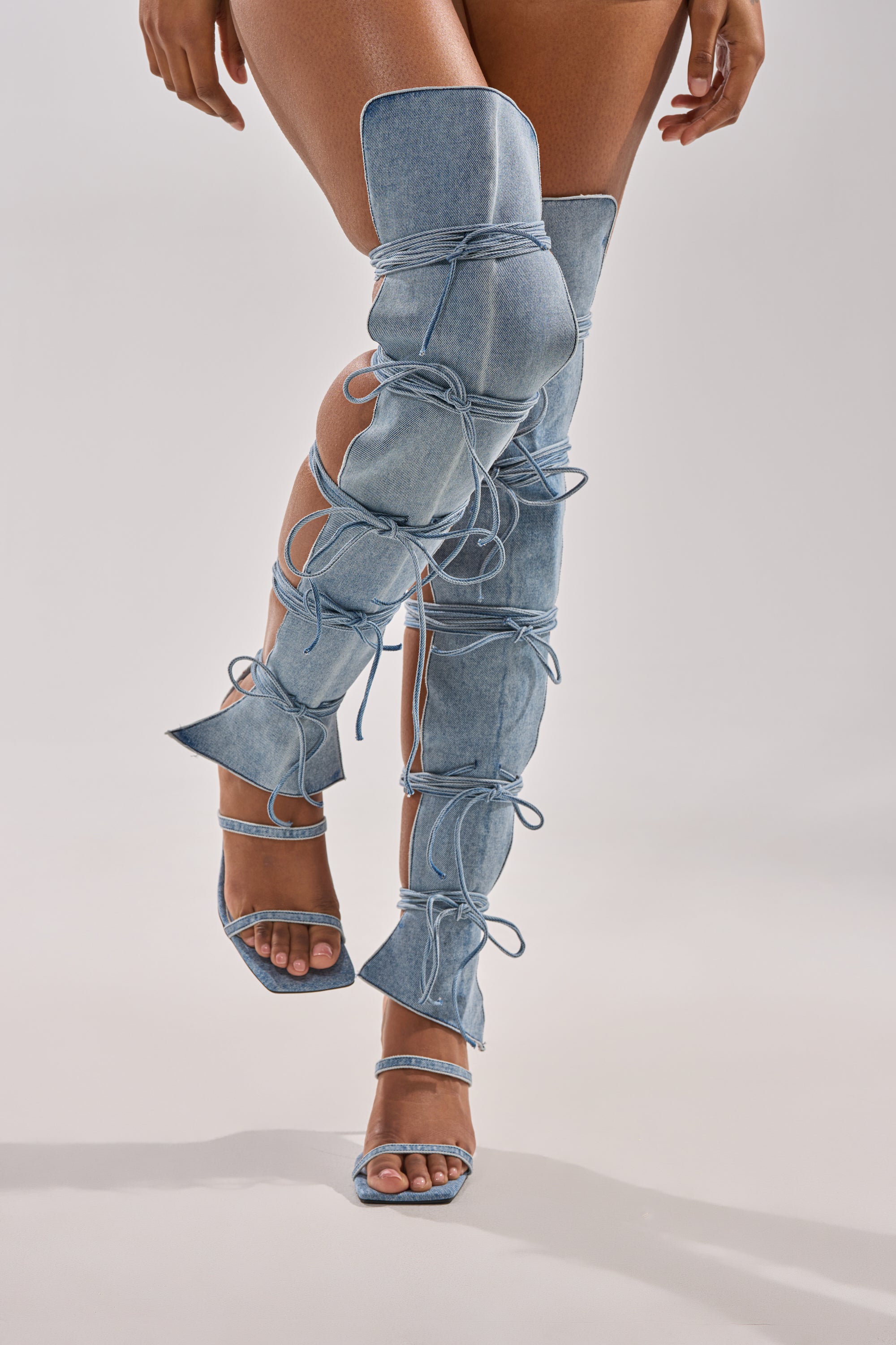 A person models the AZALEA WANG KRISTINE DENIM STILETTO GLADIATOR SANDAL—unique thigh-high, light blue denim heels with open toes, open heels, cut-out details, multiple bows, and a stylish lace-up design against a plain background.