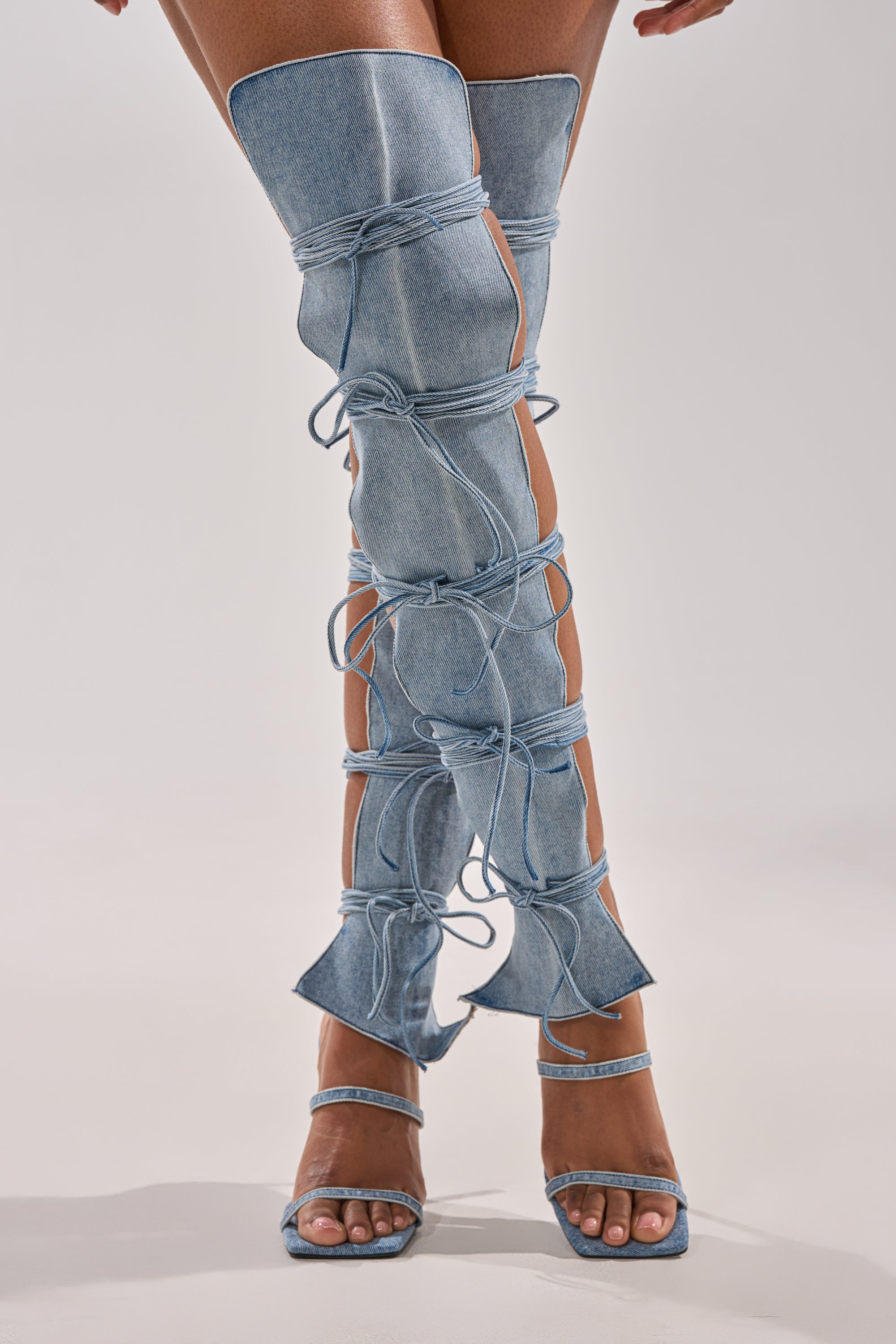 A person wears the AZALEA WANG KRISTINE DENIM STILETTO GLADIATOR SANDAL—light blue, thigh-high, open-sided denim boots with denim laces and a matching open square toe high heel.