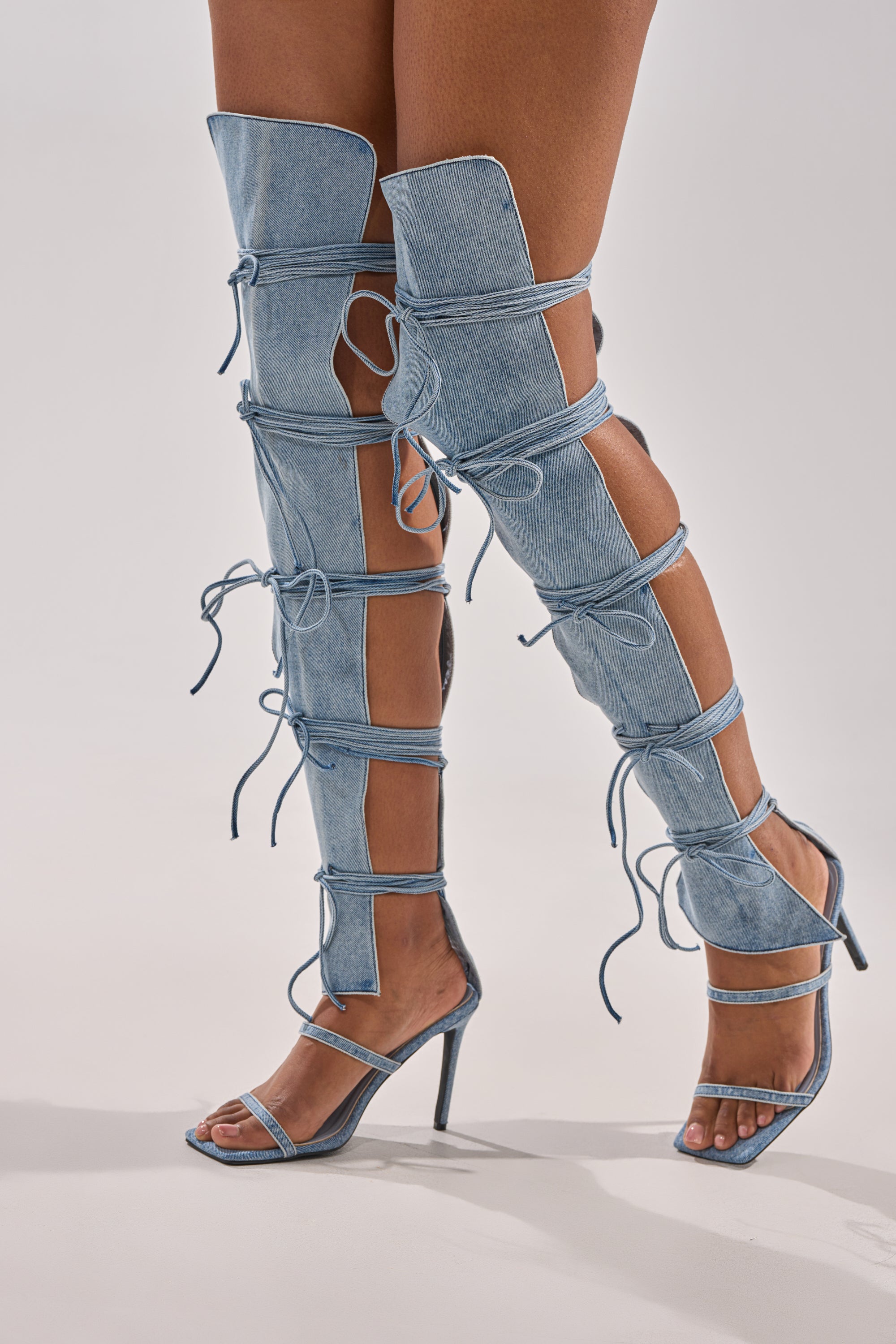 A person models the AZALEA WANG KRISTINE DENIM STILETTO GLADIATOR SANDAL—an open-toe, thigh-high denim boot featuring multiple cutouts and lace-up details—against a plain light background.