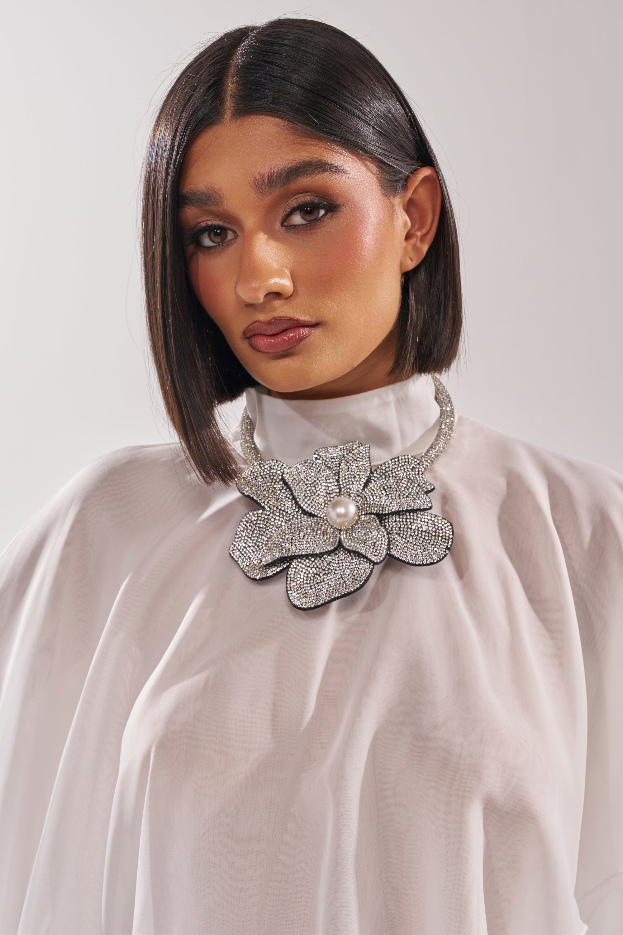 A woman with straight dark hair poses against a plain background wearing a high-neck white blouse and the HOLIDAY READY NECKLACE, a large statement piece featuring crystals and a central pearl. 1