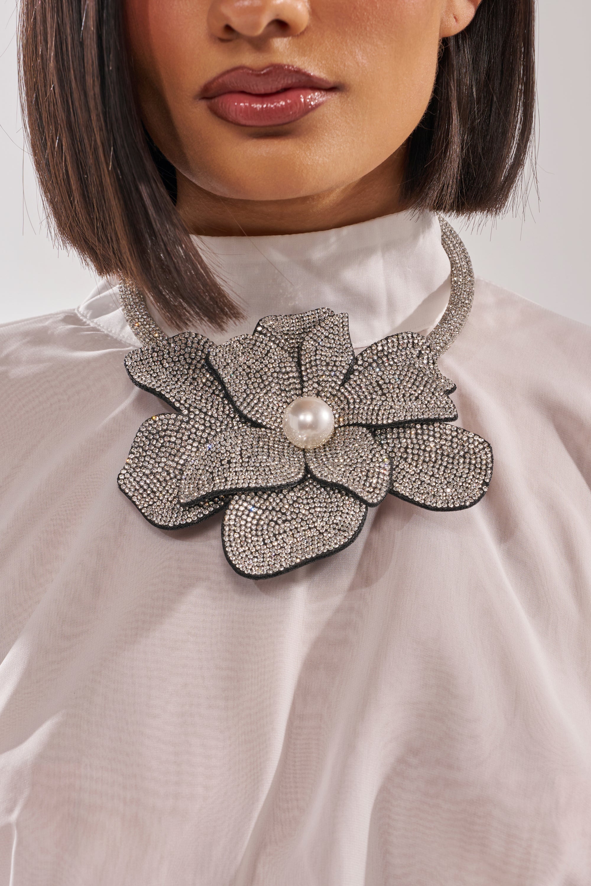 A woman wears the HOLIDAY READY NECKLACE—a pearl statement piece with a jeweled floral pendant and sparkling rhinestones—paired with a sheer white top. Only her lower face and neck are shown. 4