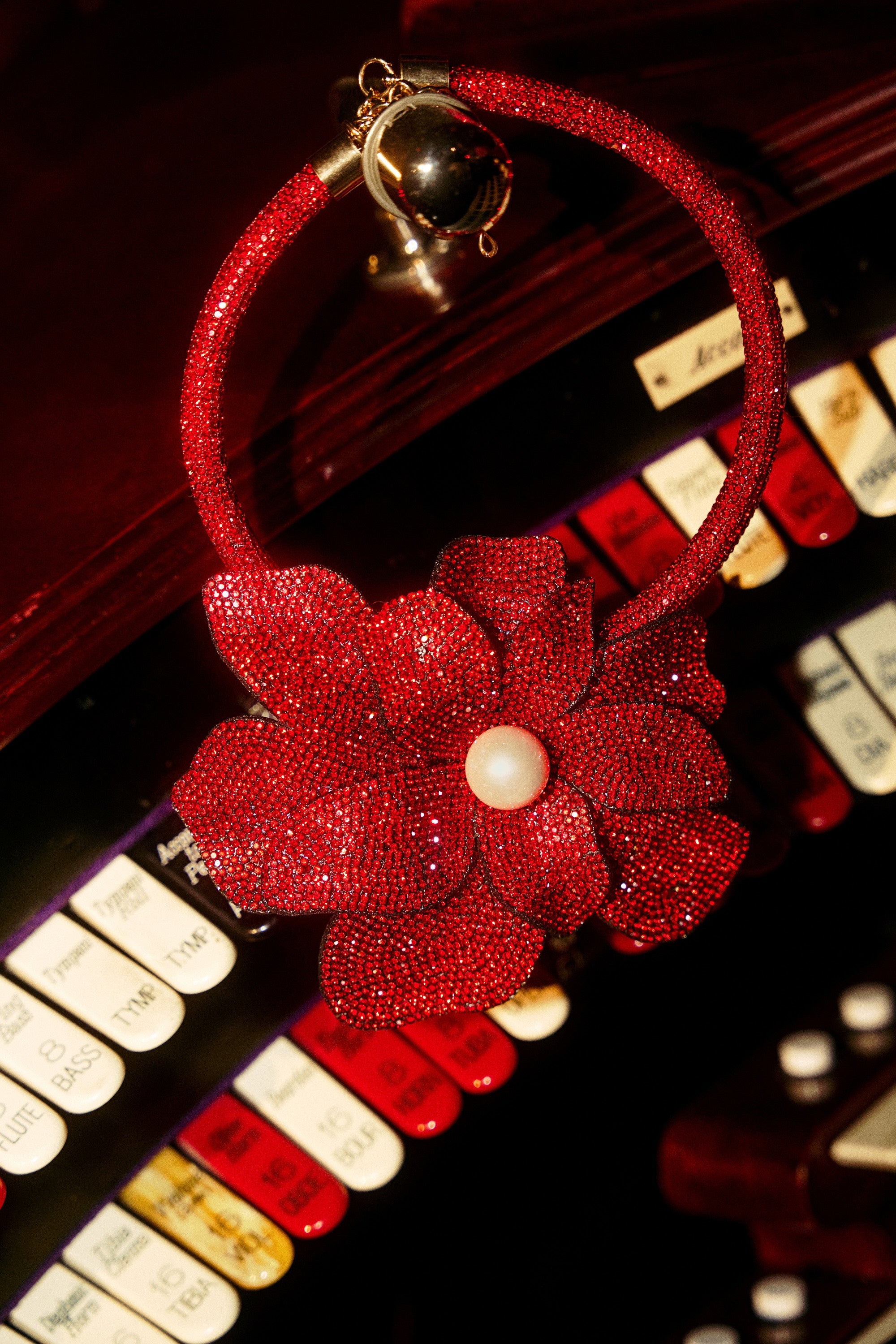 The IN BLOOM NECKLACE, featuring a sparkling red flower with a pearl center, rests on vintage organ keys next to a statement necklace; white and red labels are visible in the background. 1