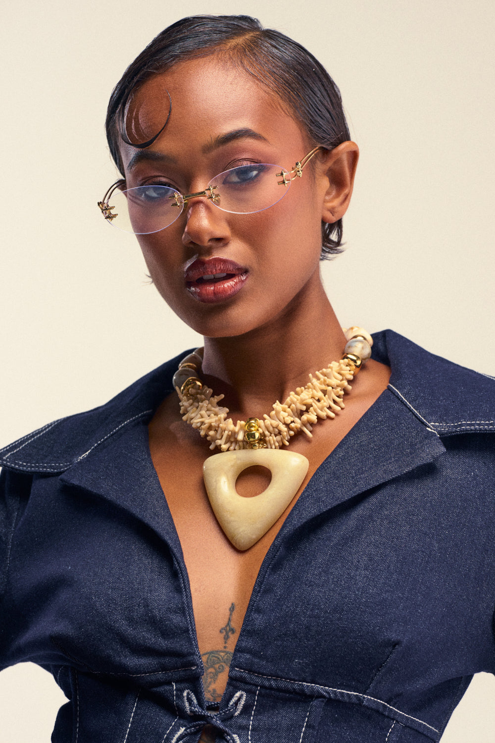 A woman in a deep blue outfit, sporting stylish glasses and short, slicked-back hair with a curl on her forehead, confidently gazes at the camera. She completes her look with the striking EARTH ANGEL NECKLACE IN WHITE against a plain background.