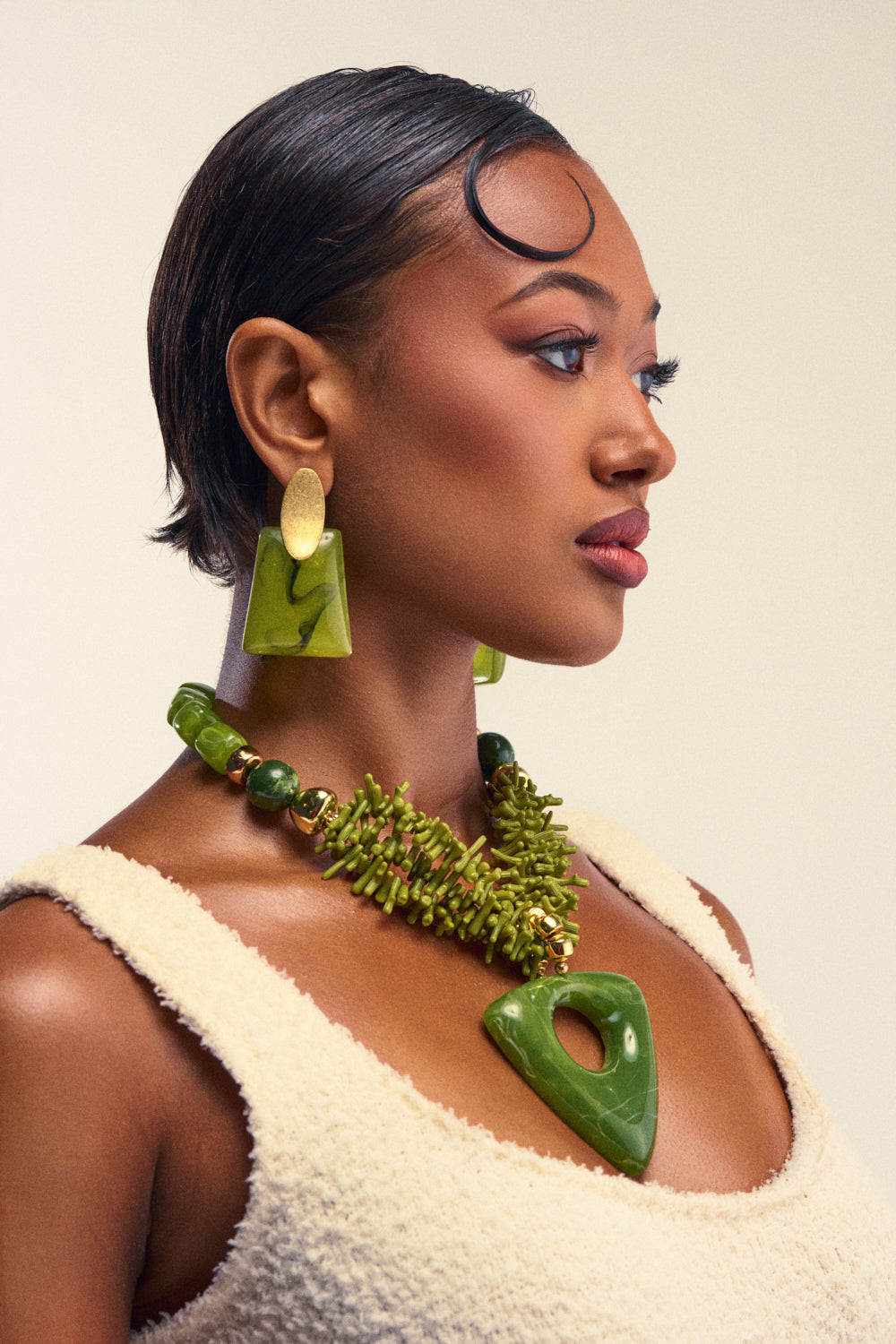 A woman with short, sleek hair wears bold green and gold jewelry, including the EARTH ANGEL NECKLACE IN GREEN as a statement pendant, paired with striking earrings and layered necklaces. Her ensemble features a textured cream top that perfectly complements her accessories as she gazes confidently to the side against a neutral background.