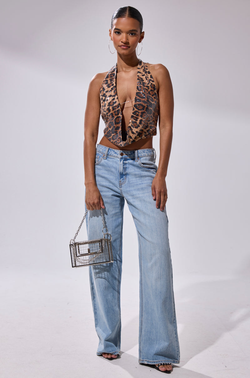 A woman faces forward in the CHEETAH GIRLY COWL NECK TOP, styled with light blue wide-leg jeans, black sandals, hoop earrings, and a small transparent box-shaped handbag against a plain light background.