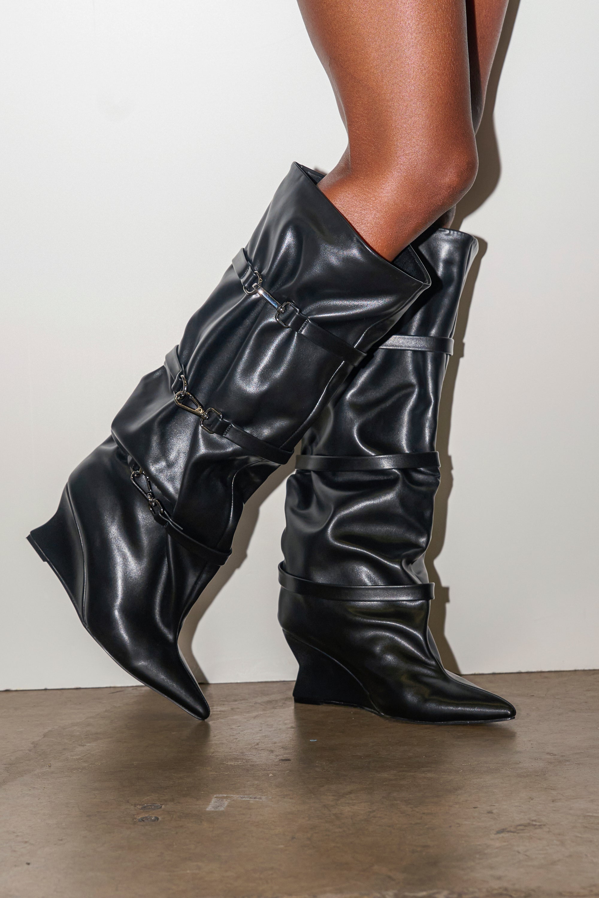 A person models the AZALEA WANG KULLERVO Black Silver Clasp Detail Wedge Boot, featuring a slouchy faux leather knee-high design with silver hardware, standing on a concrete floor against a plain white background.