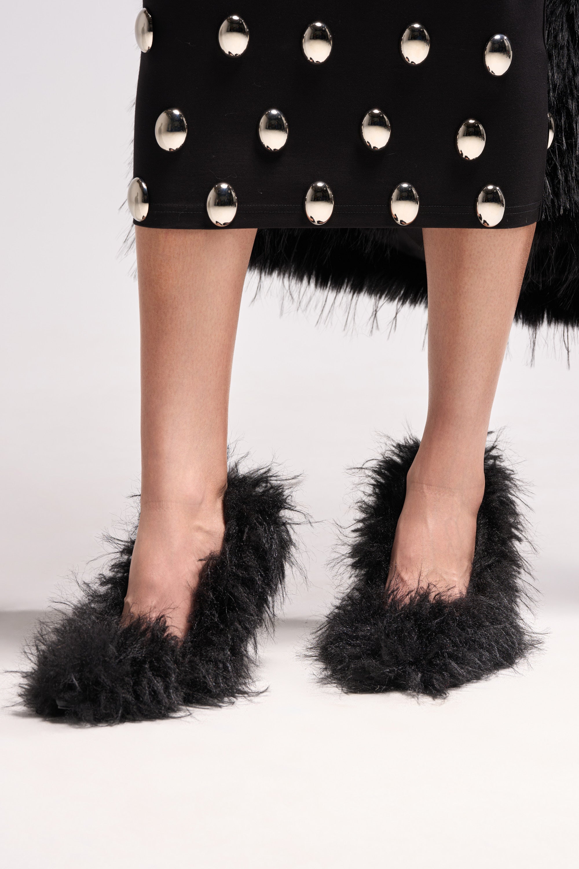 A person wears the AZALEA WANG KURT BLACK FUR EMBELLISHED PUMP with a black skirt featuring large silver studs, showing only their lower legs and skirt against a plain background.