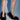 A person stands on a light blue surface with shadows, wearing only the AZALEA WANG KURT BLACK FUR EMBELLISHED PUMP—black faux fur stilettos. Only their legs and feet are visible.