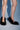 A person stands on a light blue surface with shadows, wearing only the AZALEA WANG KURT BLACK FUR EMBELLISHED PUMP—black faux fur stilettos. Only their legs and feet are visible.