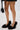 A person in a pinstriped skirt with a black handbag stands on a white surface, showcasing the AZALEA WANG KURT BLACK FUR EMBELLISHED PUMP—black high heels with fluffy faux fur and an elegant pointed toe.