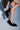 A person in a black pinstripe skirt slips on the AZALEA WANG KURT BLACK FUR EMBELLISHED PUMP, one leg raised and hands adjusting the shoe, against a soft blue background.