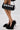 Wearing AZALEA WANG KURT BLACK FUR EMBELLISHED PUMPS, a person stands with crossed legs, holding a black handbag decorated with five wristwatches. The scene features a plain white background.