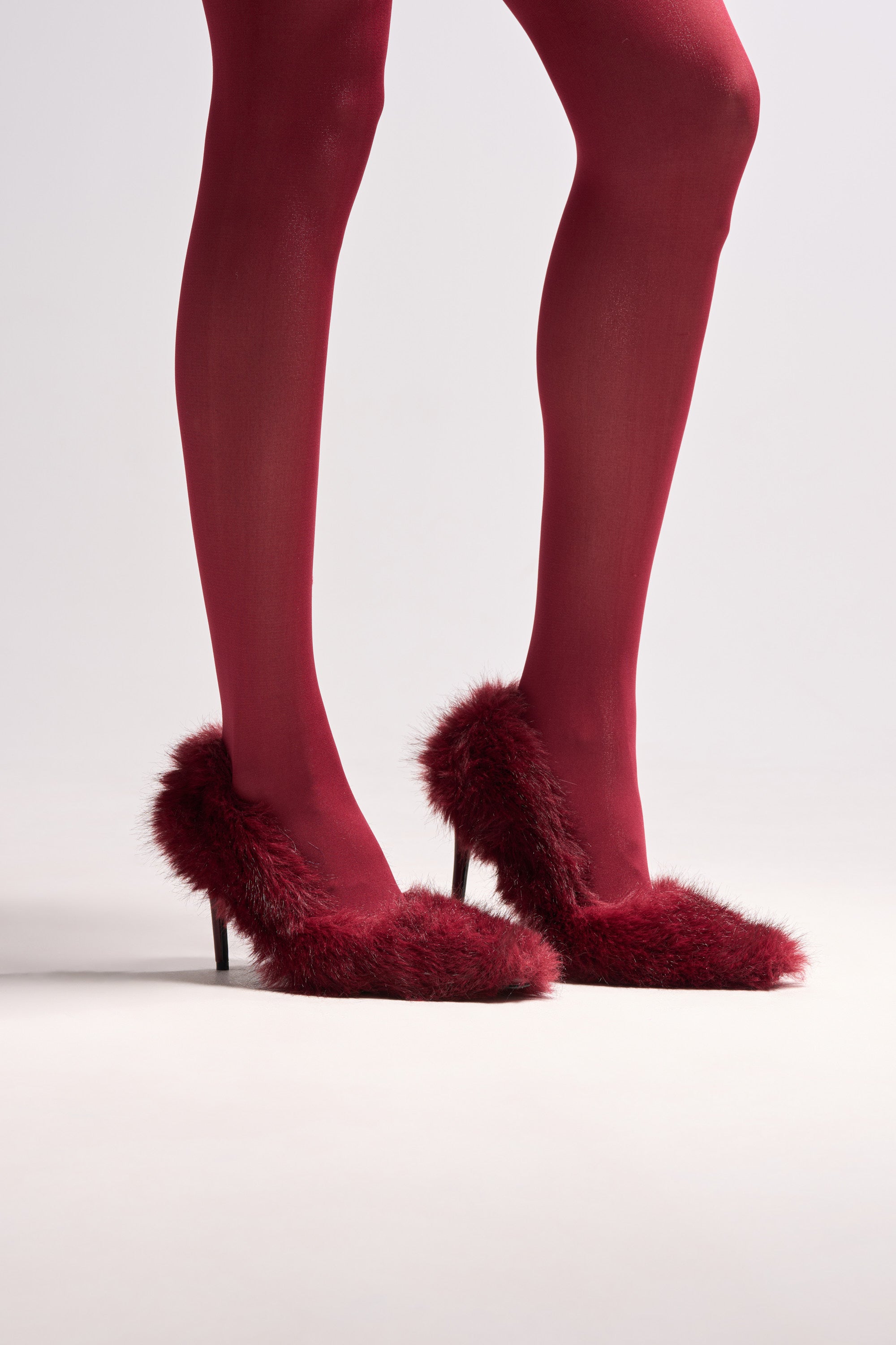 A person wears sheer burgundy tights and the AZALEA WANG KURT BURGUNDY FUR EMBELLISHED PUMP, featuring a faux fur upper and stiletto heel, standing on a light-colored surface.