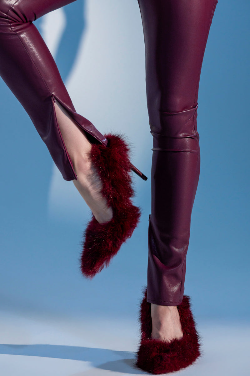 A person wearing burgundy faux leather pants with ankle zippers and AZALEA WANG KURT BURGUNDY FUR EMBELLISHED PUMPS stands against a light blue background.