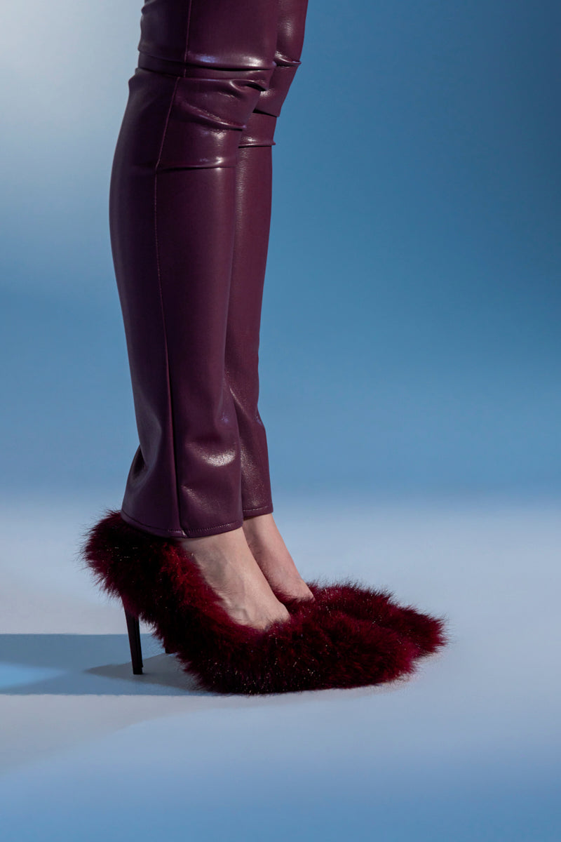 A person in shiny burgundy pants and AZALEA WANG KURT BURGUNDY FUR EMBELLISHED PUMP stilettos stands on a blue and white background.