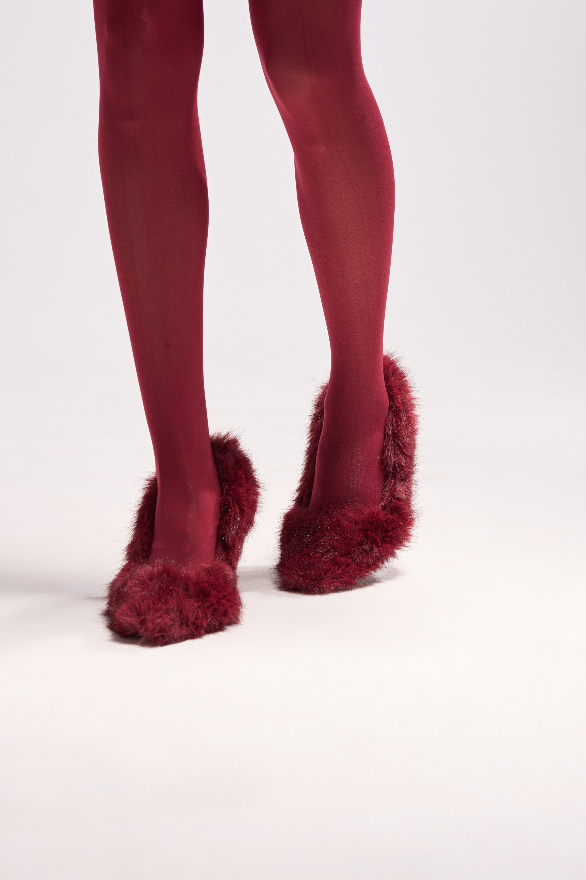 A person in opaque red tights and AZALEA WANG KURT BURGUNDY FUR EMBELLISHED PUMPS with stiletto heels stands on a plain, light background. The photo is cropped at the knees, emphasizing the bold legs and striking footwear.