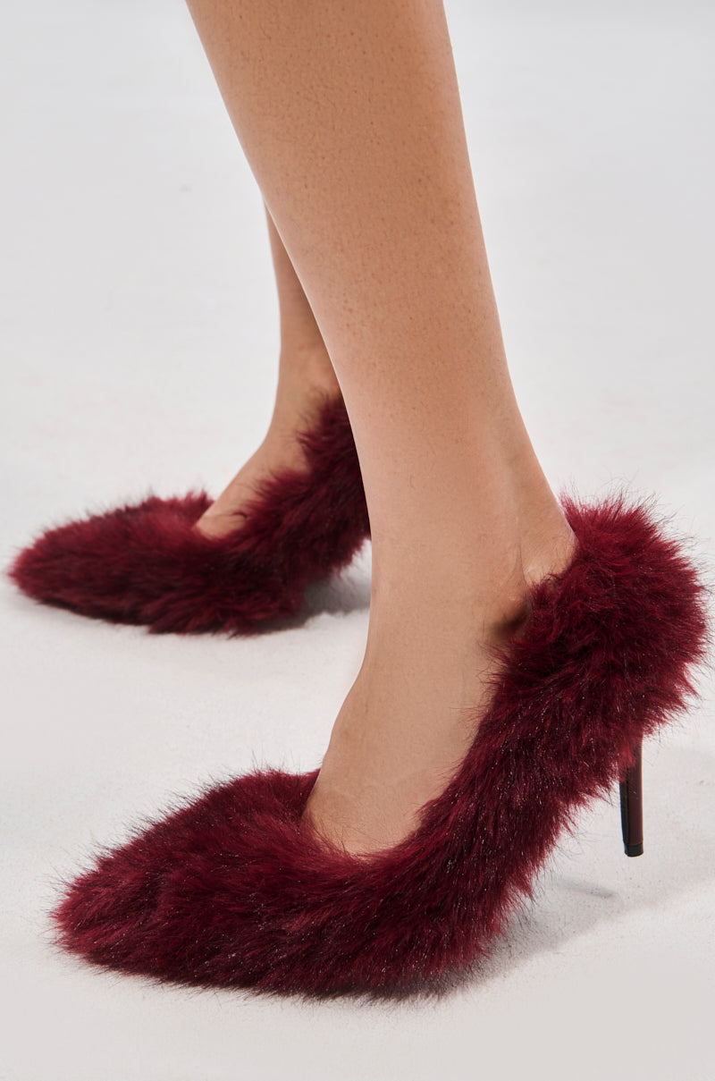 A person wears the AZALEA WANG KURT BURGUNDY FUR EMBELLISHED PUMP, featuring a faux fur upper, pointed toes, and striking stiletto heels, while standing on a light-colored surface.